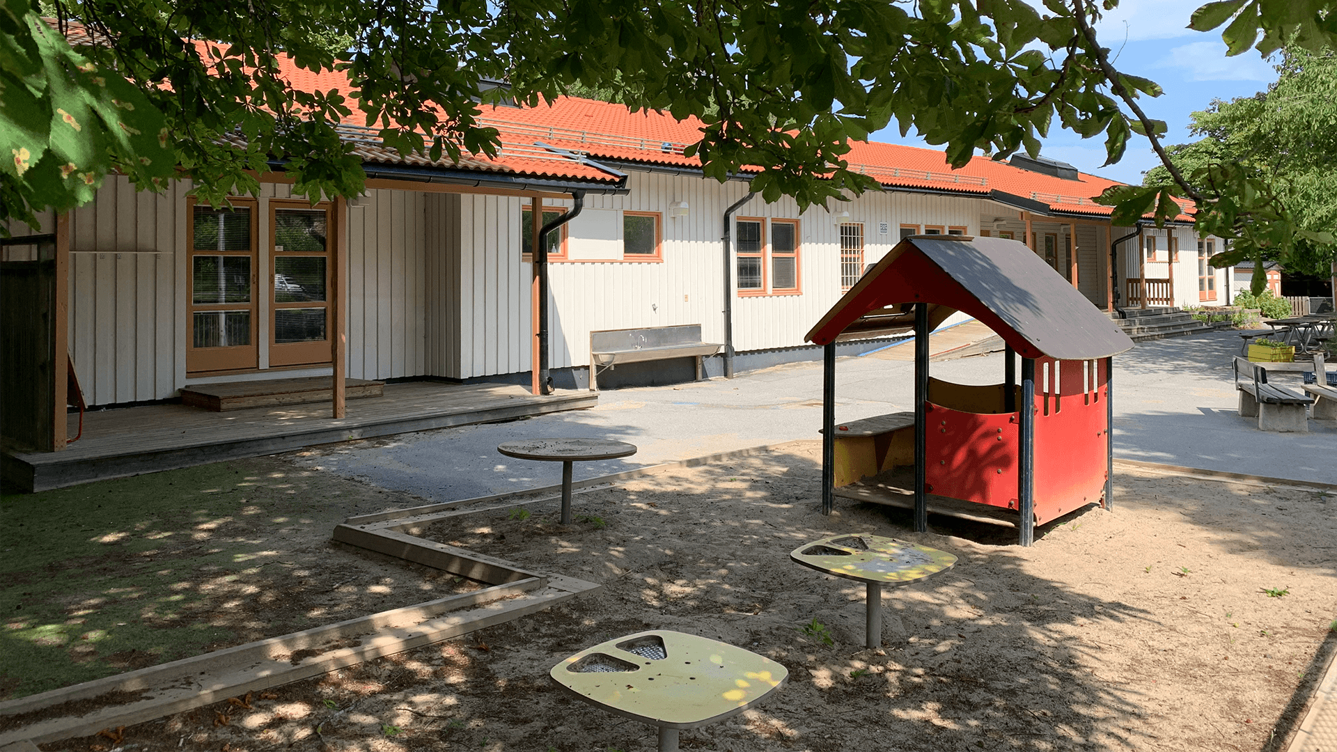 Lummig gård med träd, lekredskap och gräsmatta framför ett vitt enplanshus i trä.