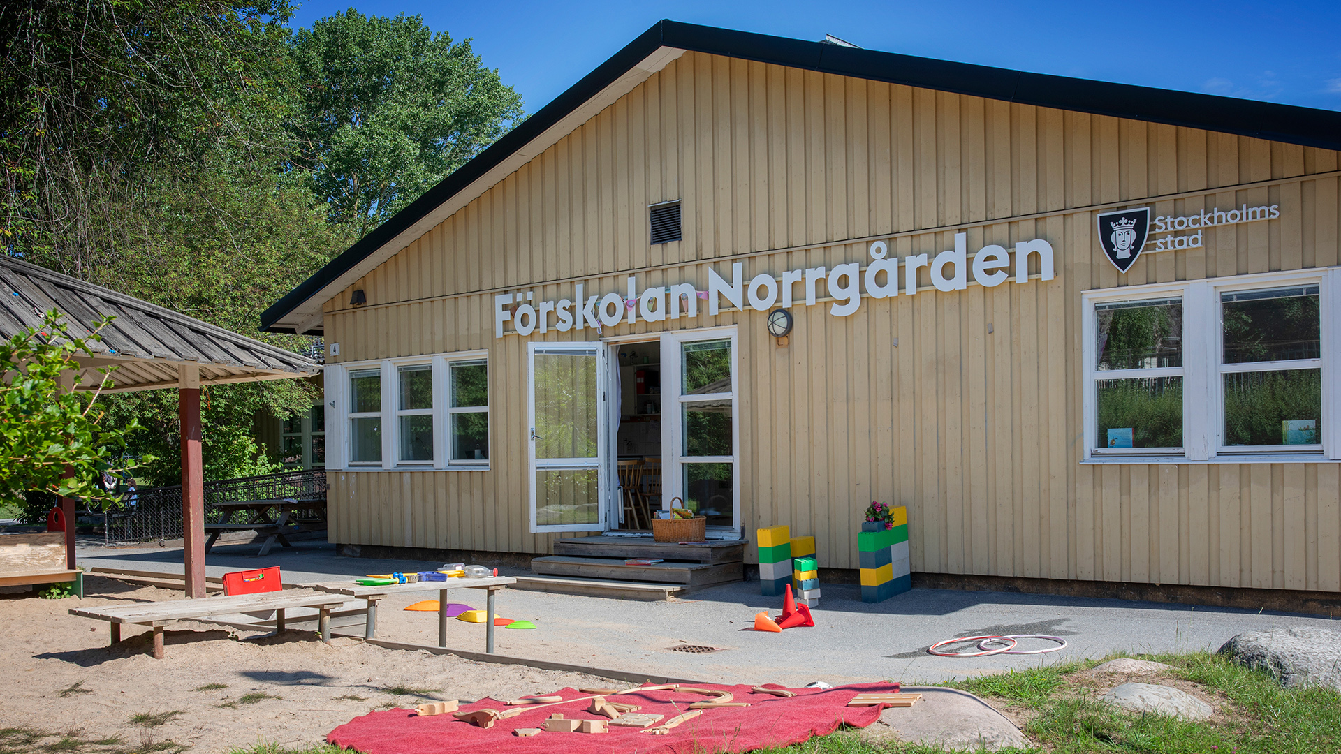 Förskolegård med träd, gräs och en stor sandlåda. I övrigt asfalt med rockringar, byggklossar och andra leksaker.