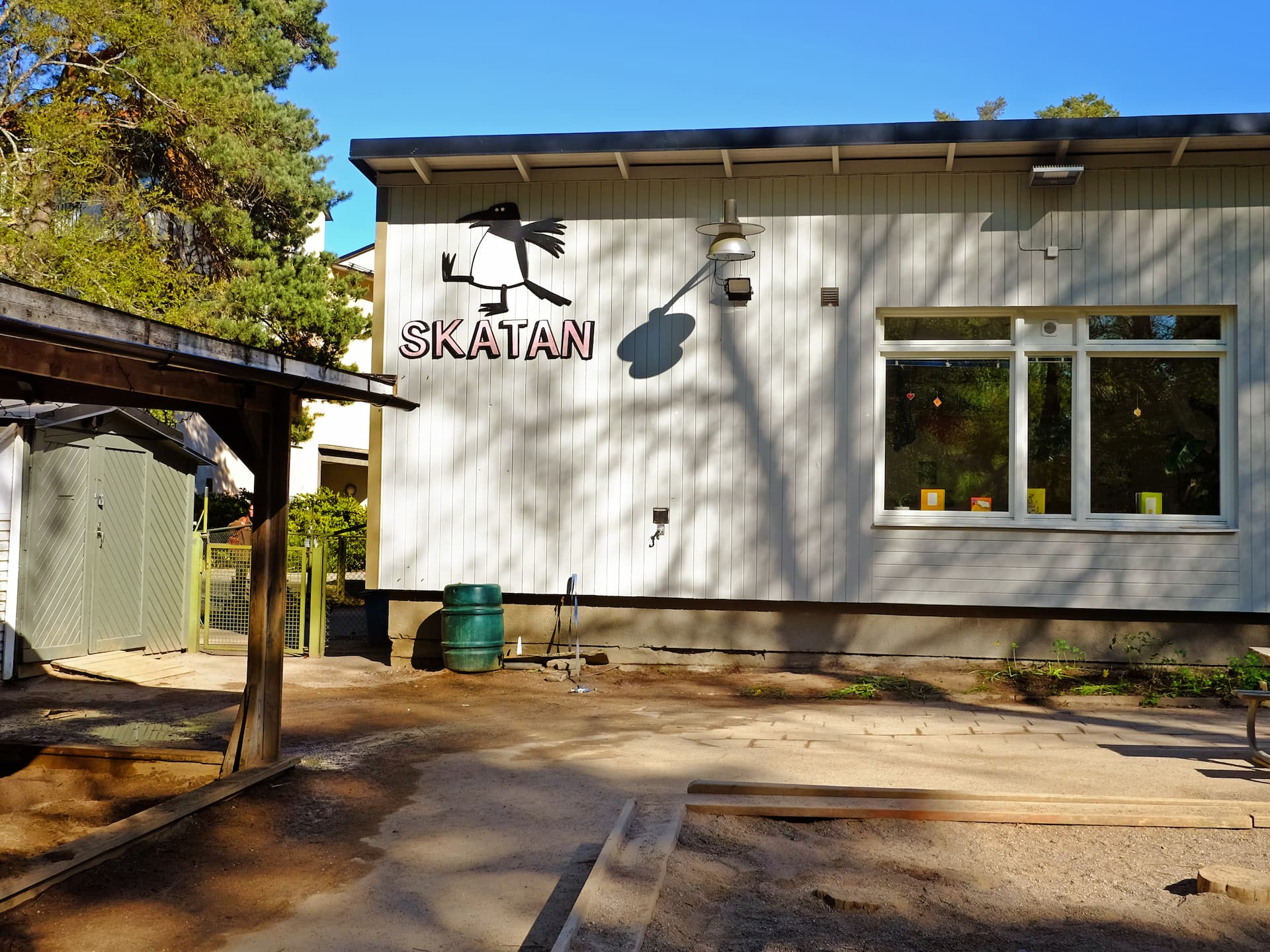 Vit byggnad med skylten ”Skatan” och fågellogga i ett soligt skogsområde, med sandlåda och gård i förgrunden.