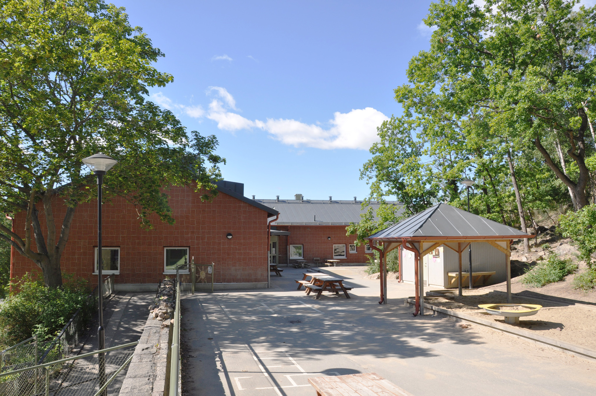 Låg tegelbyggnad med stor gård med grönskande träd, bänkar och pergola.