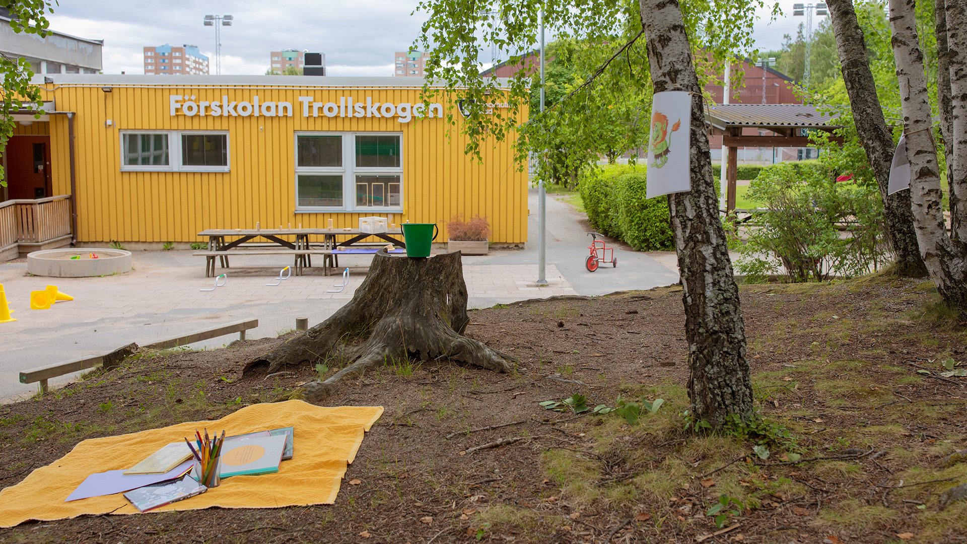 Förskolegård med träd, kuperad terräng och buskar. I övrigt asfalt med bord, trehjulingar och andra leksaker samt sandlåda.