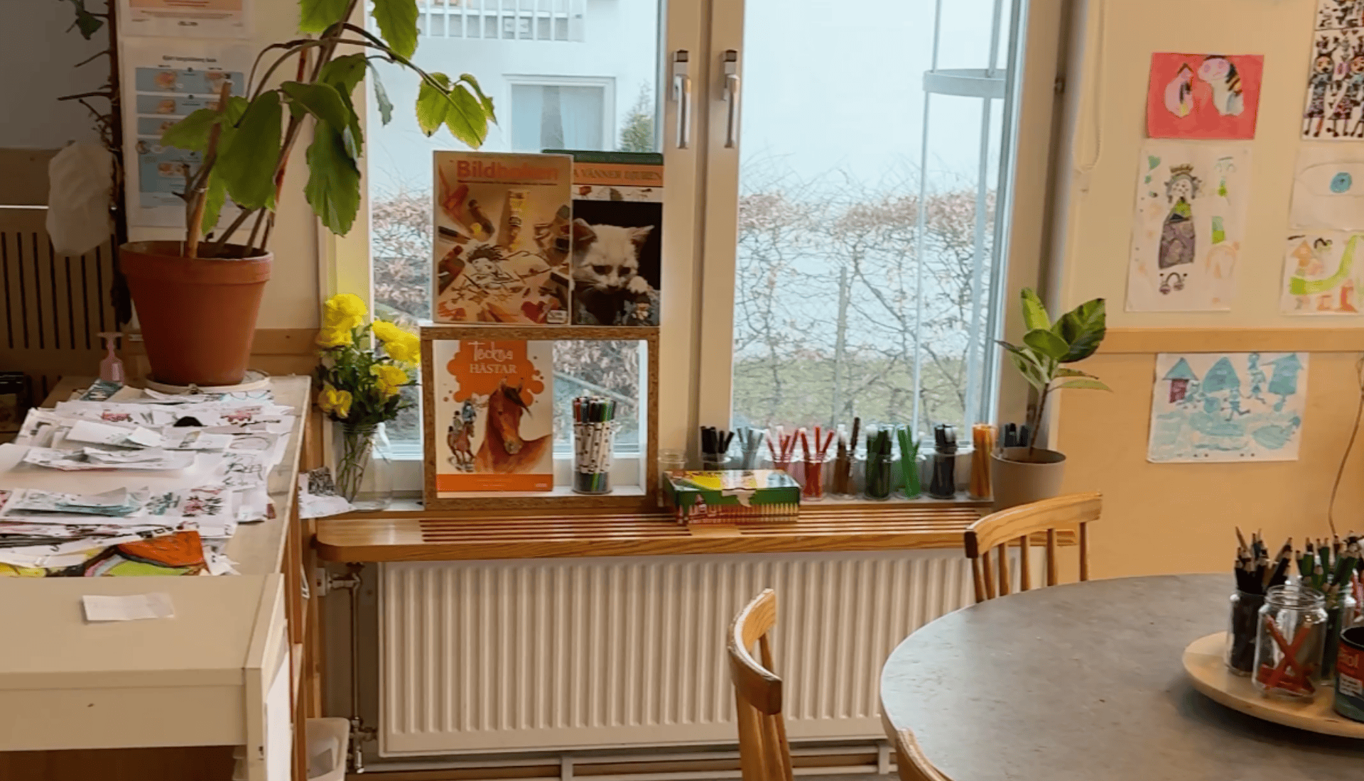 Inomhusvy med fönsterbräda fylld med böcker, pennor och blommor. Bord med papper och konstmaterial.