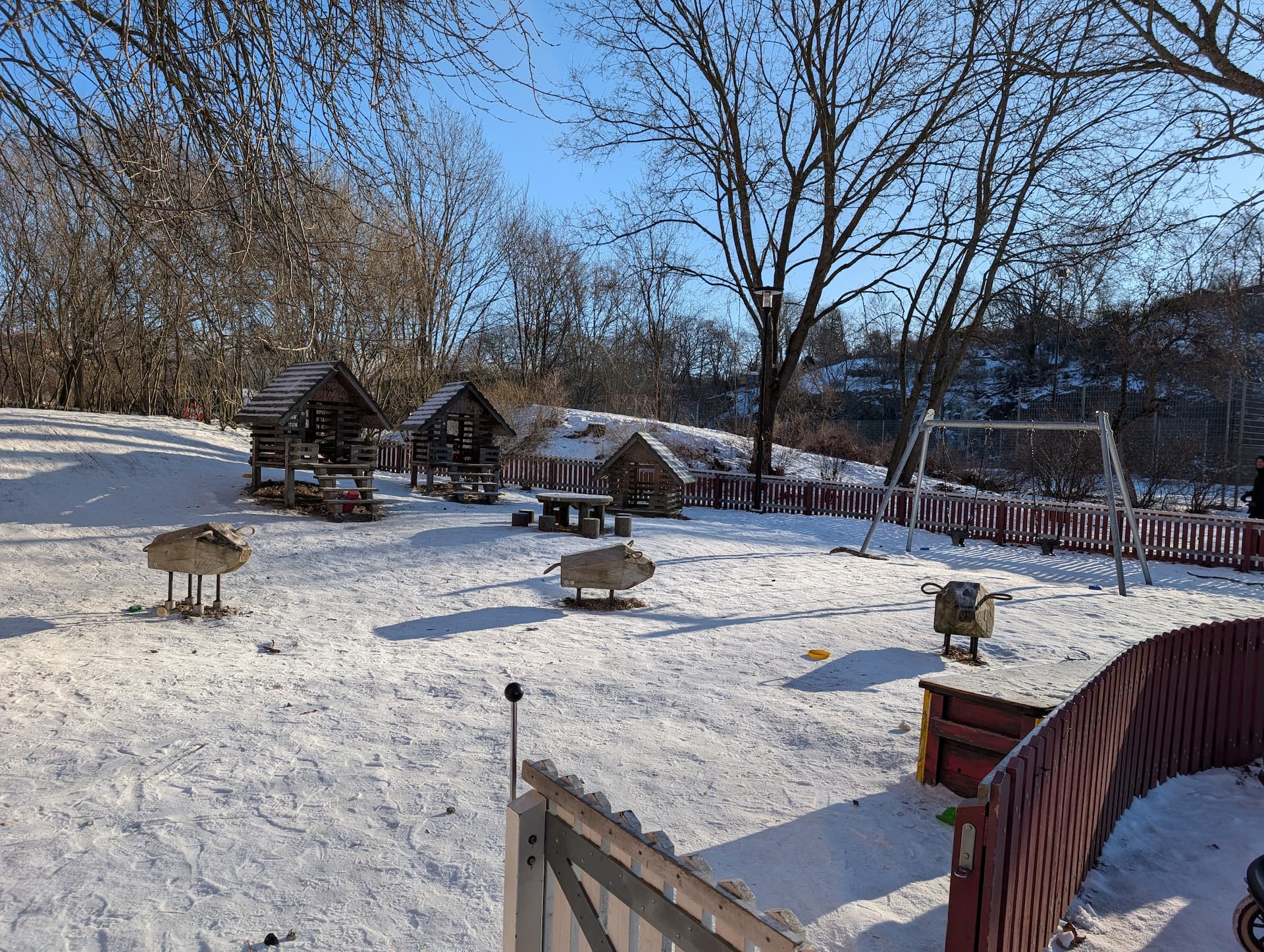 Snötäckt lekplats med små träkojor, gungor och lekdjur, omgiven av träd i vinterlandskap.
