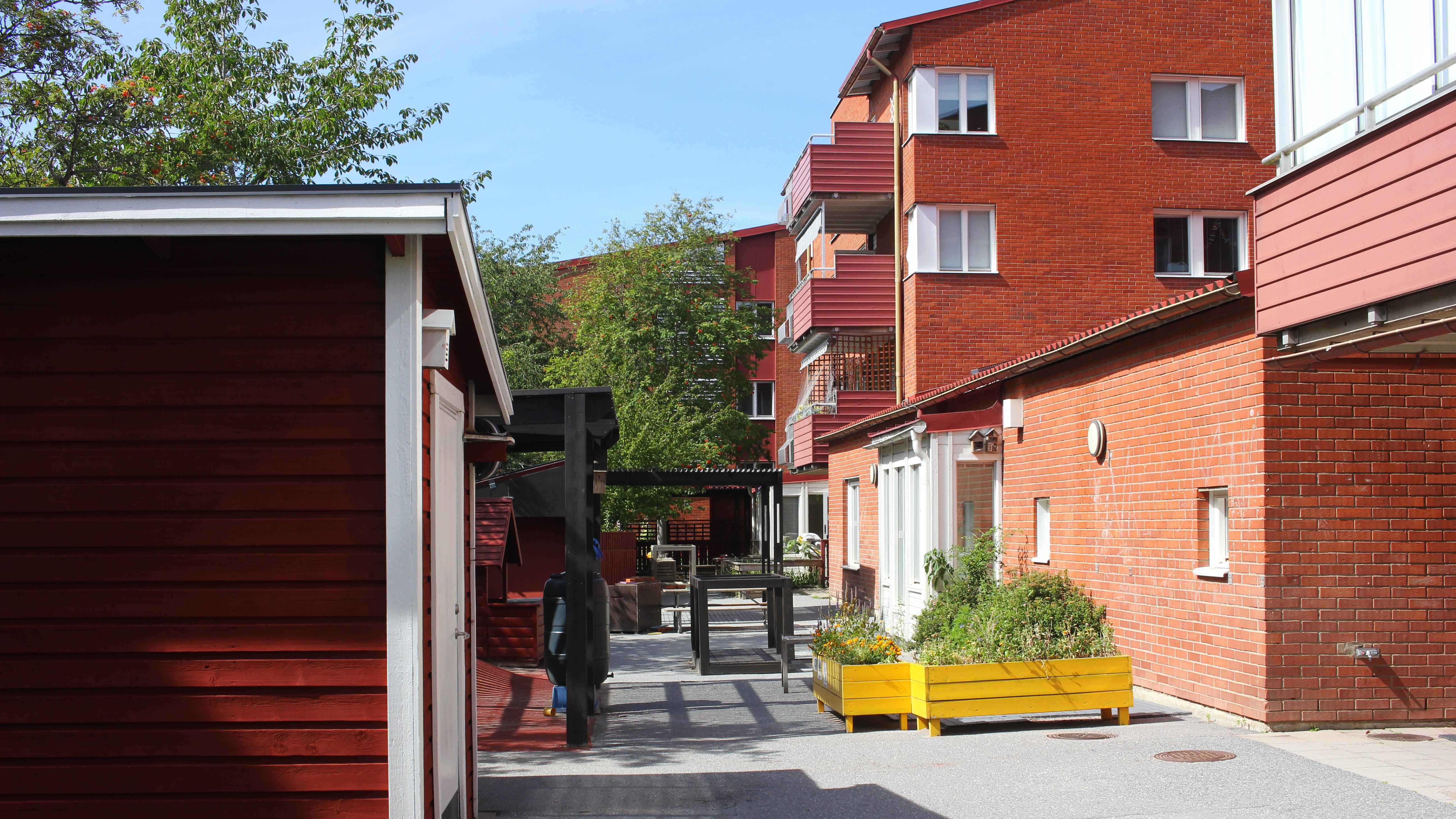 Ett flerfamiljshus i rött tegel och en asfalterad gård med ett rött förråd och en gul planteringslåda med blommor. En lekyta med ett rött lekhus skymtas bakom förrådet.
