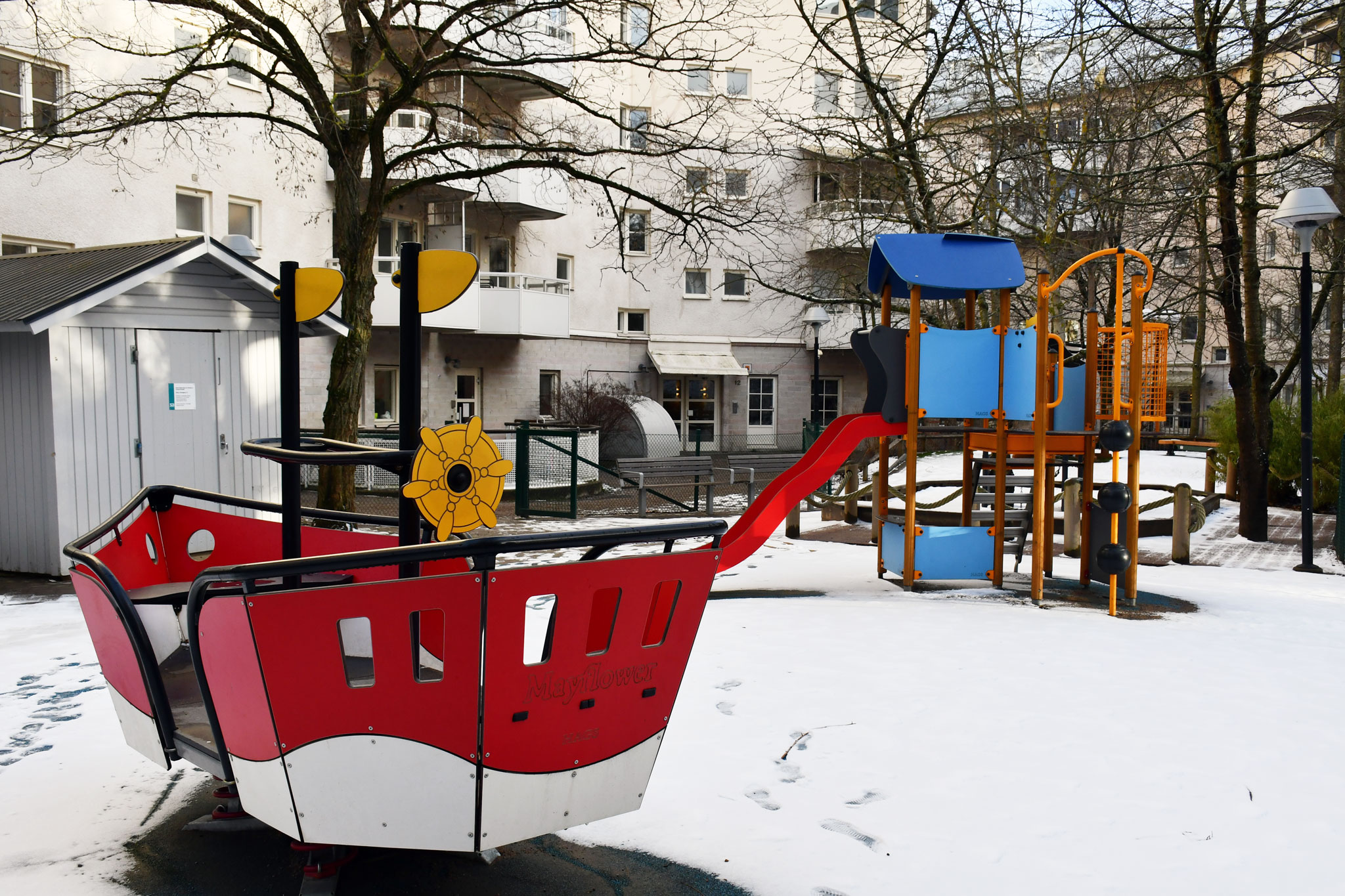 En snöig ljus innergård omgiven av flervåningshus, på gården finns färgglada lekredskap i form av en båt samt en hög rutschkana
