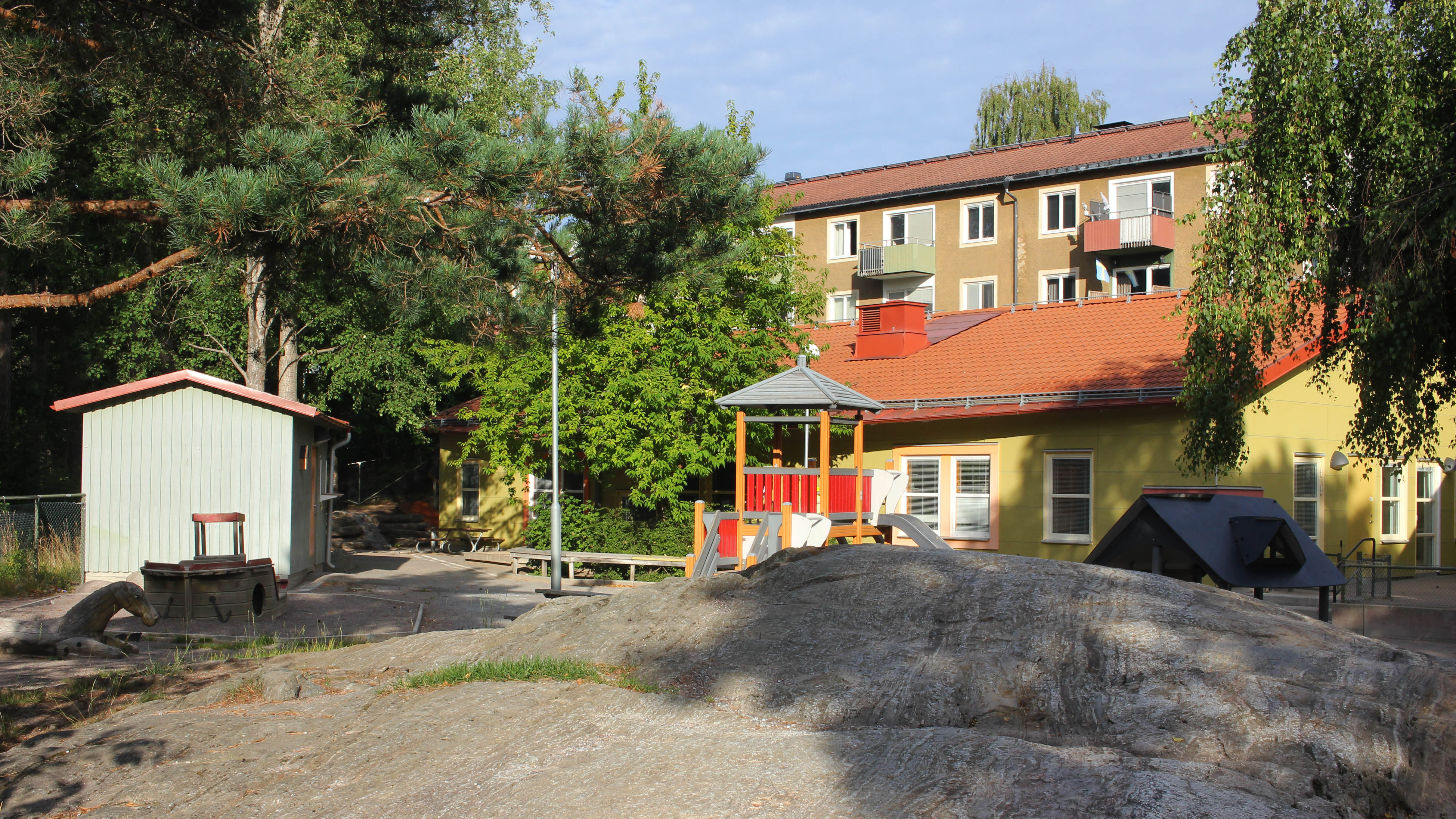 Förskolan Ankaret, ett gult enplanshus med en gård. På gården syns en gul och röd klätterställning och småhus, berg och träd. Bakom gården och förskolan synd ett flerfamiljshus i brunputsad fasad.