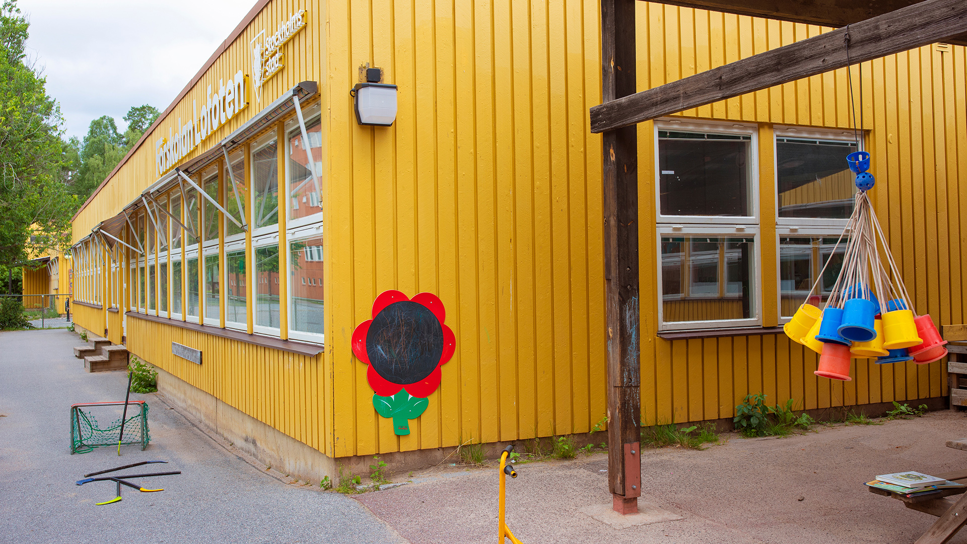 Förskolegård med träd och buskar. I övrigt asfalt med bord, sparkcykel, bandymål och bandyklubbor samt andra leksaker.