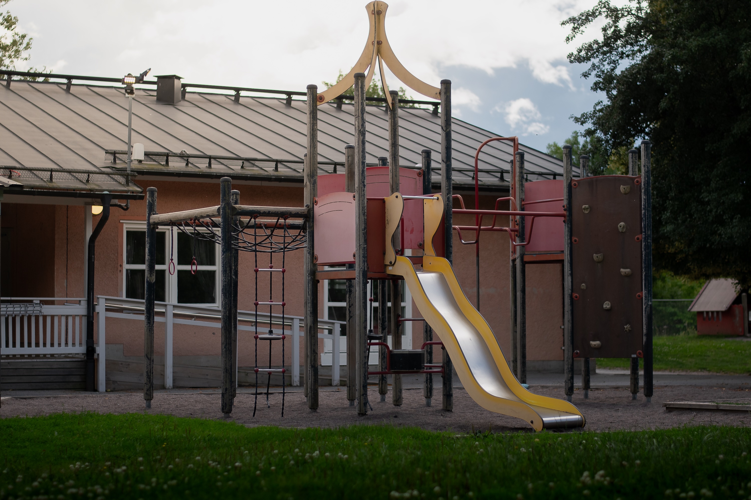 Förskolans lekplats med klätterställning och gul rutschkana framför byggnaden, omgiven av gräs och träd.