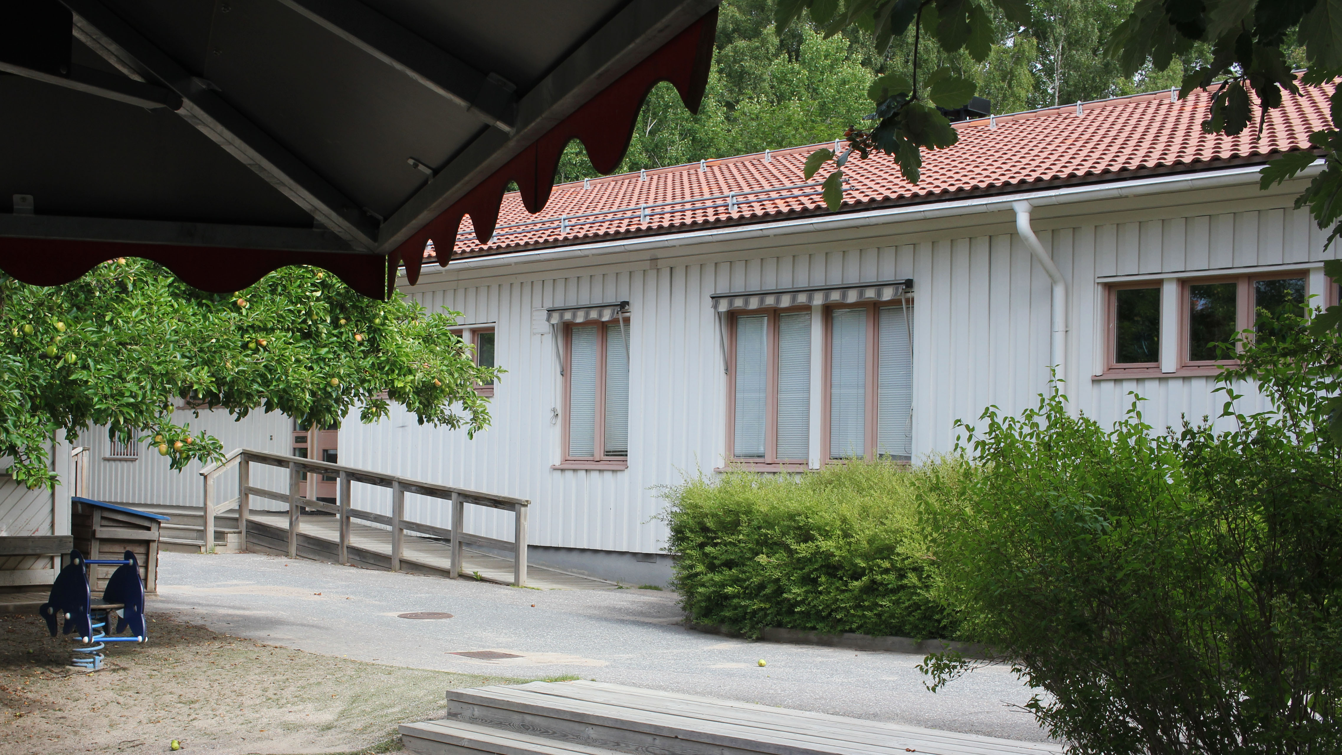 Ett enplanshus i vitt trä, rosa fönsterkarmar och ljusrött tegeltak. På gården finns ett äppelträd och framför ingången till huset finns en sandlåda och en blå gunga och ett parasol i trä. En trappa och buskar syns intill sandlådan.