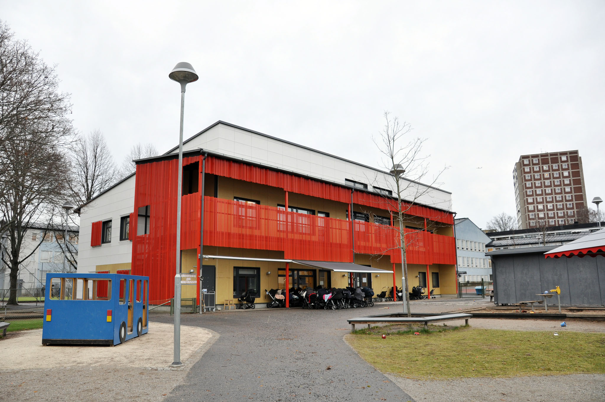 En modern byggnad i två våningar med stor balkong,sandlåda och lekredskap i form av en liten buss på gården framför.