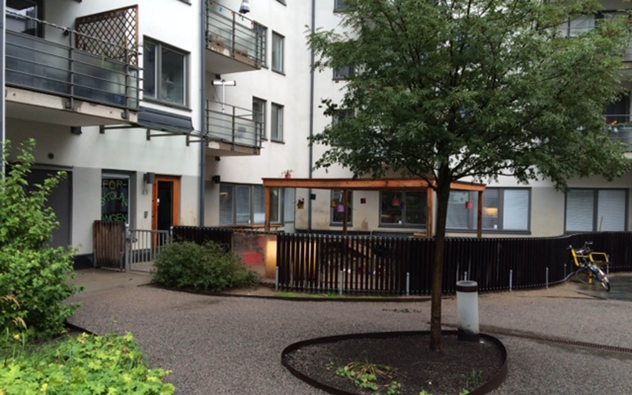 Förskolans entré med utemiljö och ett träd i förgrunden. Vitt flerbostadshus i bakgrunden.