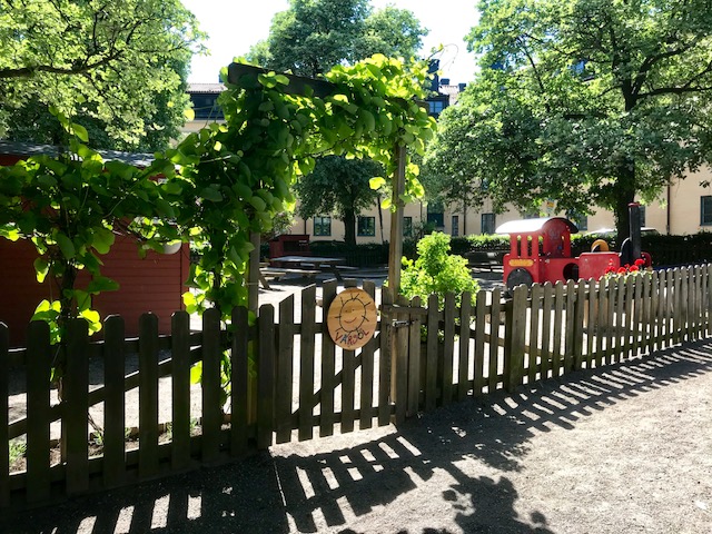 ingång till förskolans gård, grind, grönskande träd och buskar