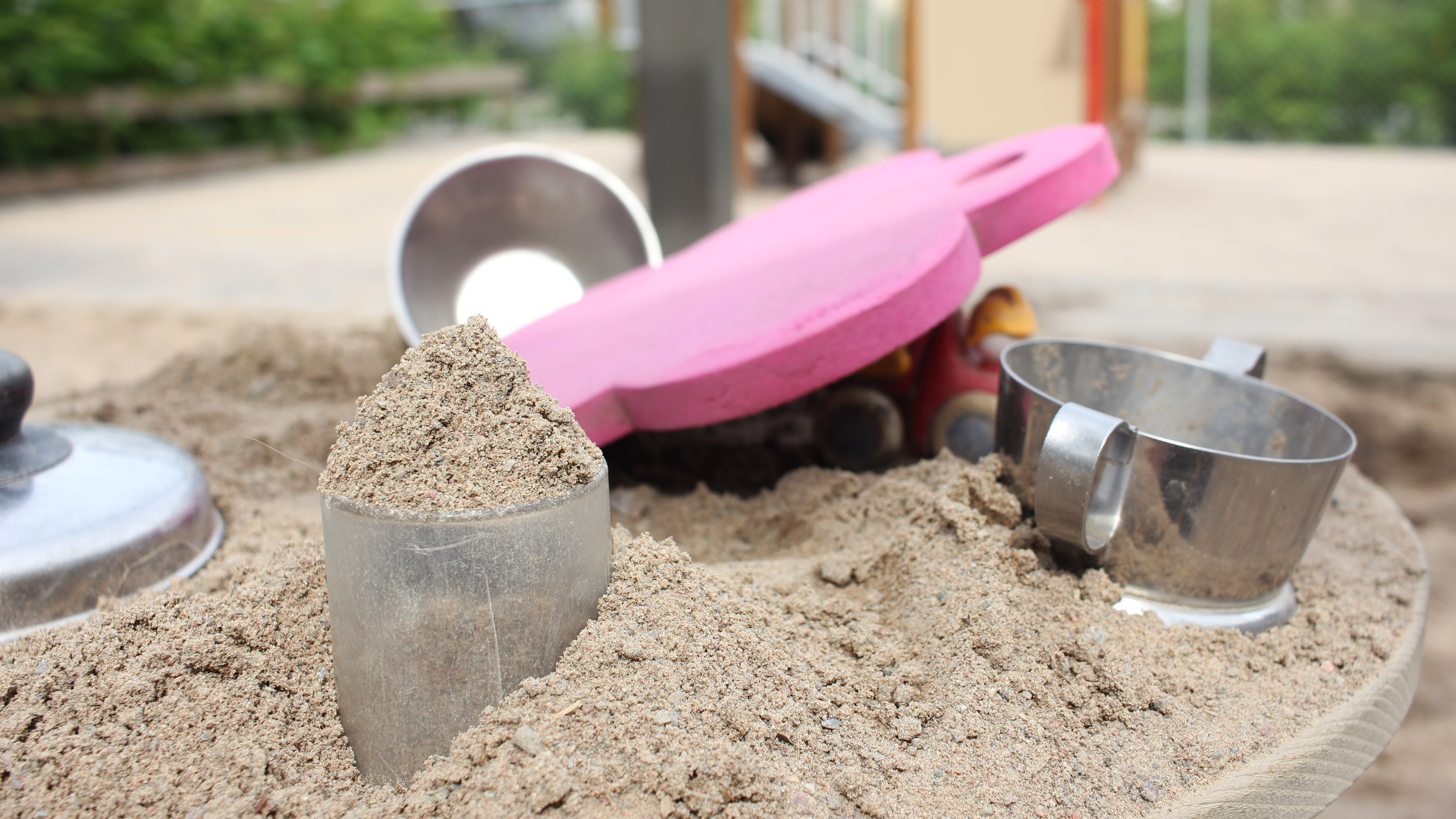 Sandleksaker i rostfritt stål och rosa ligger i en sandlåda. Bakom skymtas en klätterställning.
