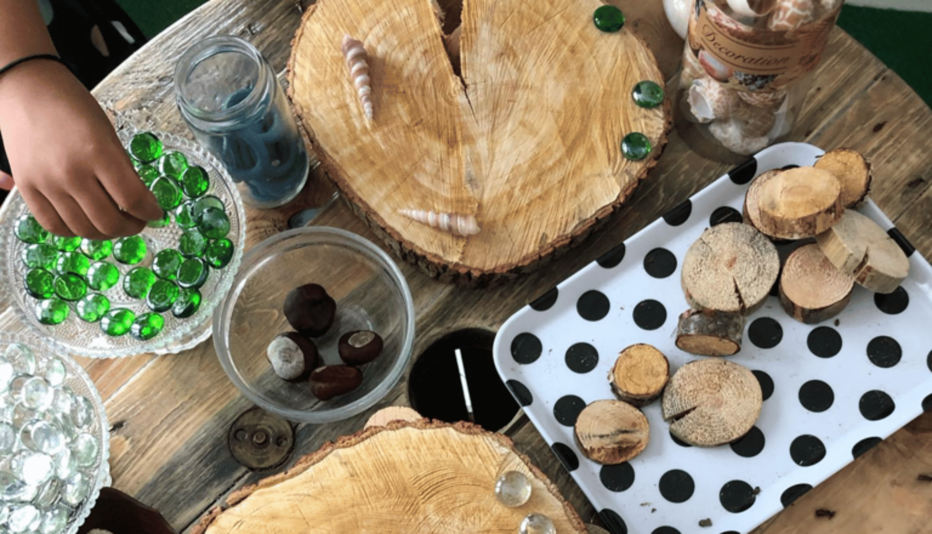 En hand som placerar gröna glasstenar på träbitar och dekorationer som snäckor och kastanjer på ett bord i en förskolemiljö.