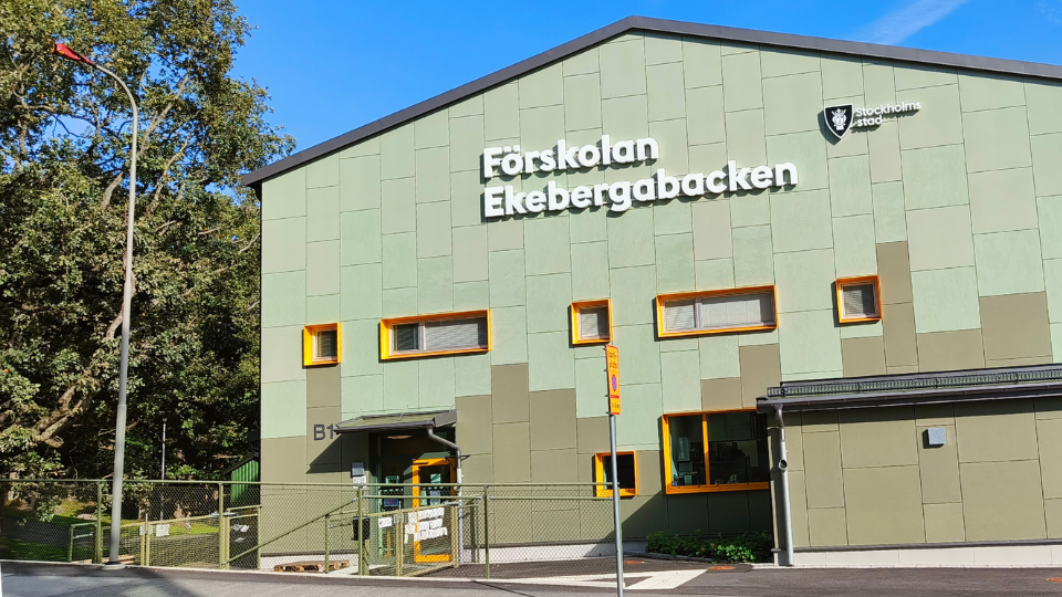 Fasaden på förskolan. Väggarna är ljust turkosa med texten "Förskolan Ekebergabacken" på. Till höger ser man träd och växtlighet från förskolans utegård. Foto. 
