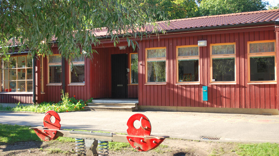 Gunga i förgrunden och förskolan i bakgrunden. Röd träpanel, enplans förskola.