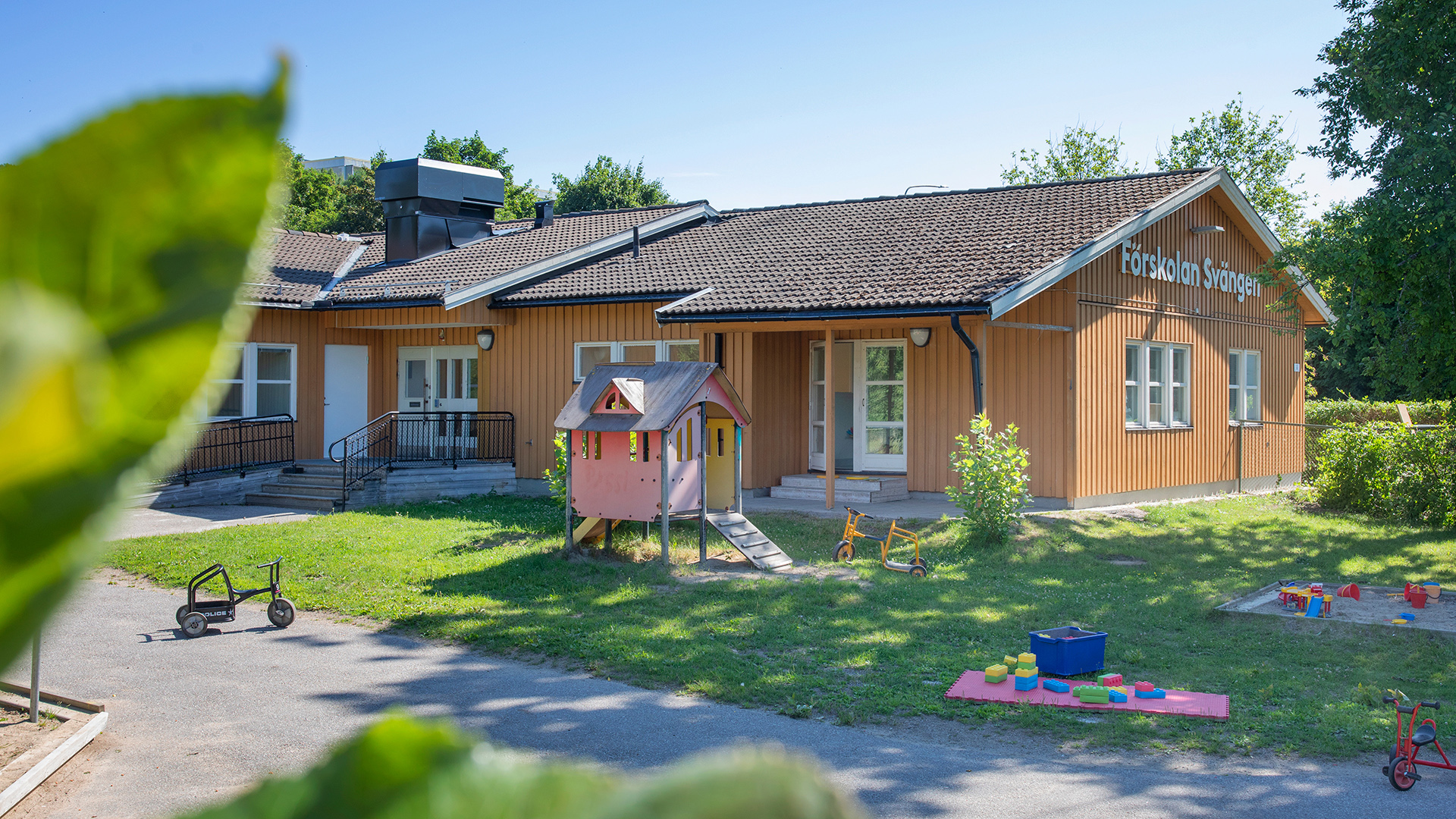 Förskolegård med gräs, buskar, träd och asfalt, samt klätterborg, sandlådor, trehjulingar och andra leksaker.