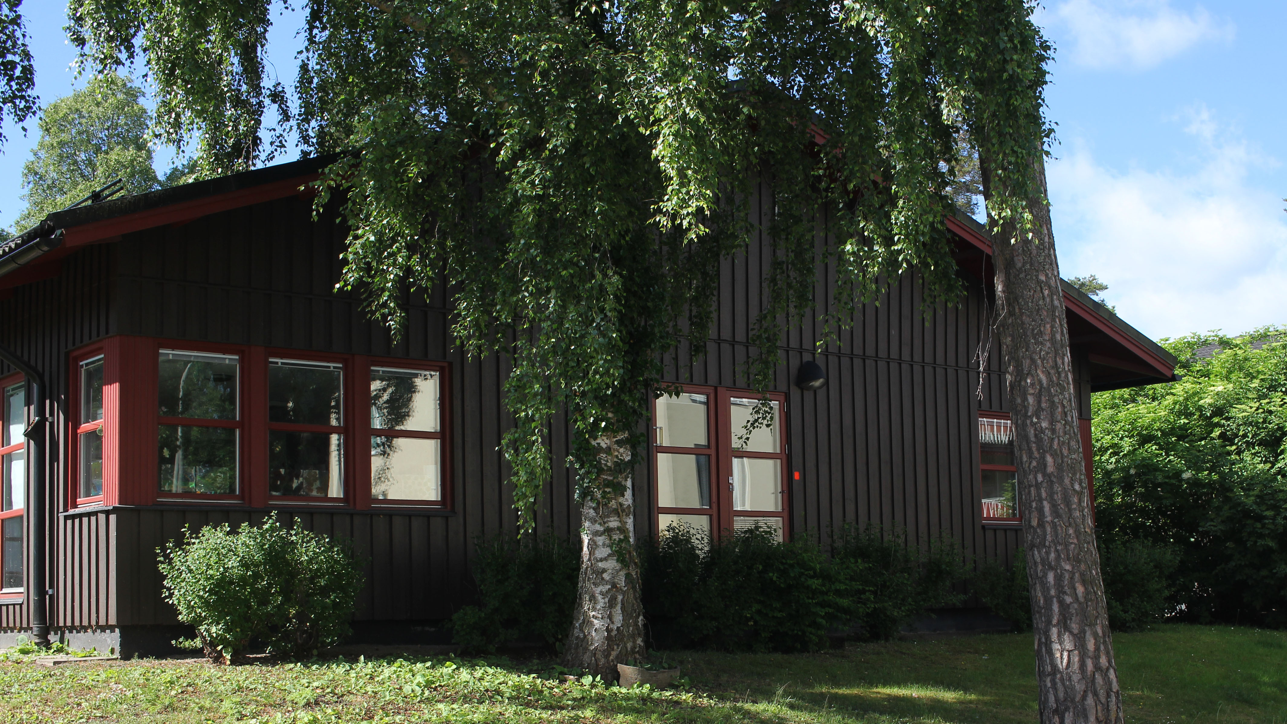 Ett brunt trähus i ett plan med röda fönsterkarmar intill gröna buskar och lövträd. Framför syns en gräsmatta.
