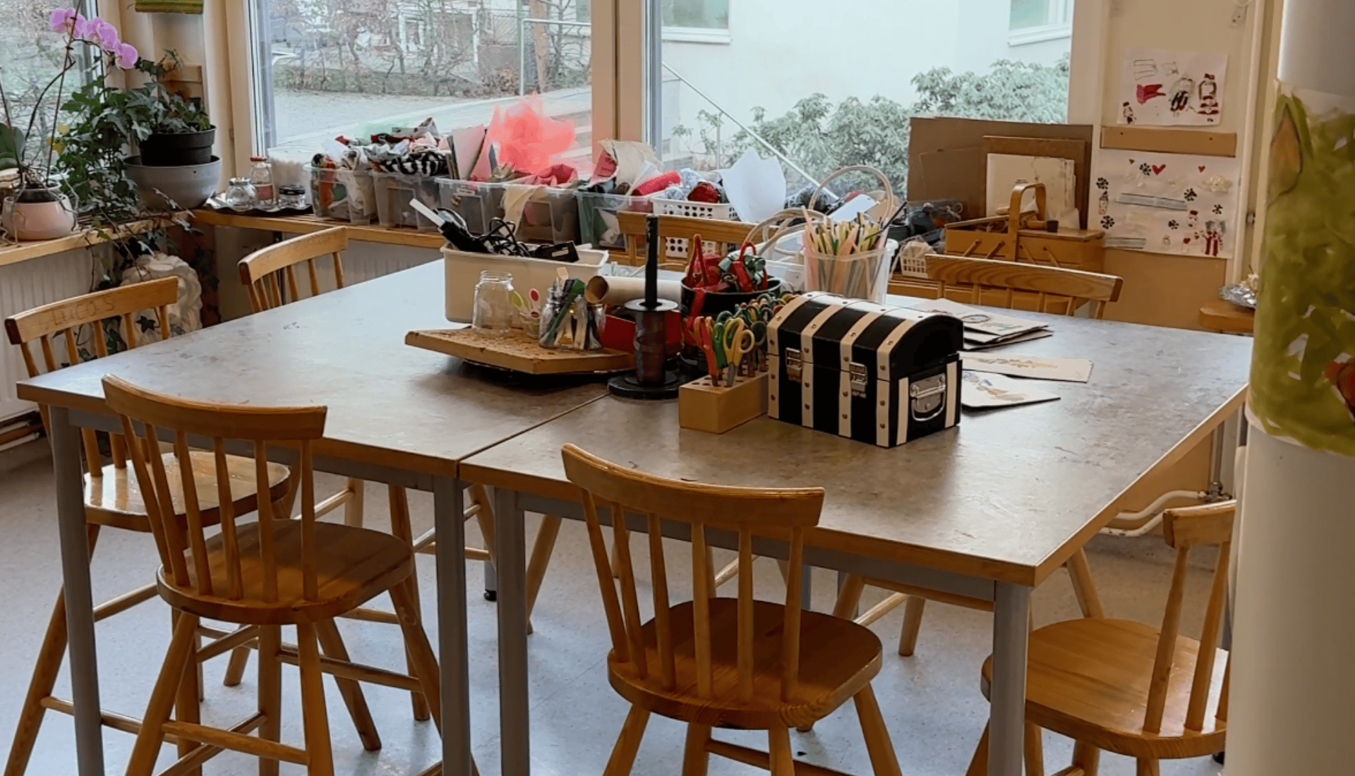 Inomhus i ett förskoleklassrum med ett stort bord, stolar och pysselmaterial på bordet samt hyllor.