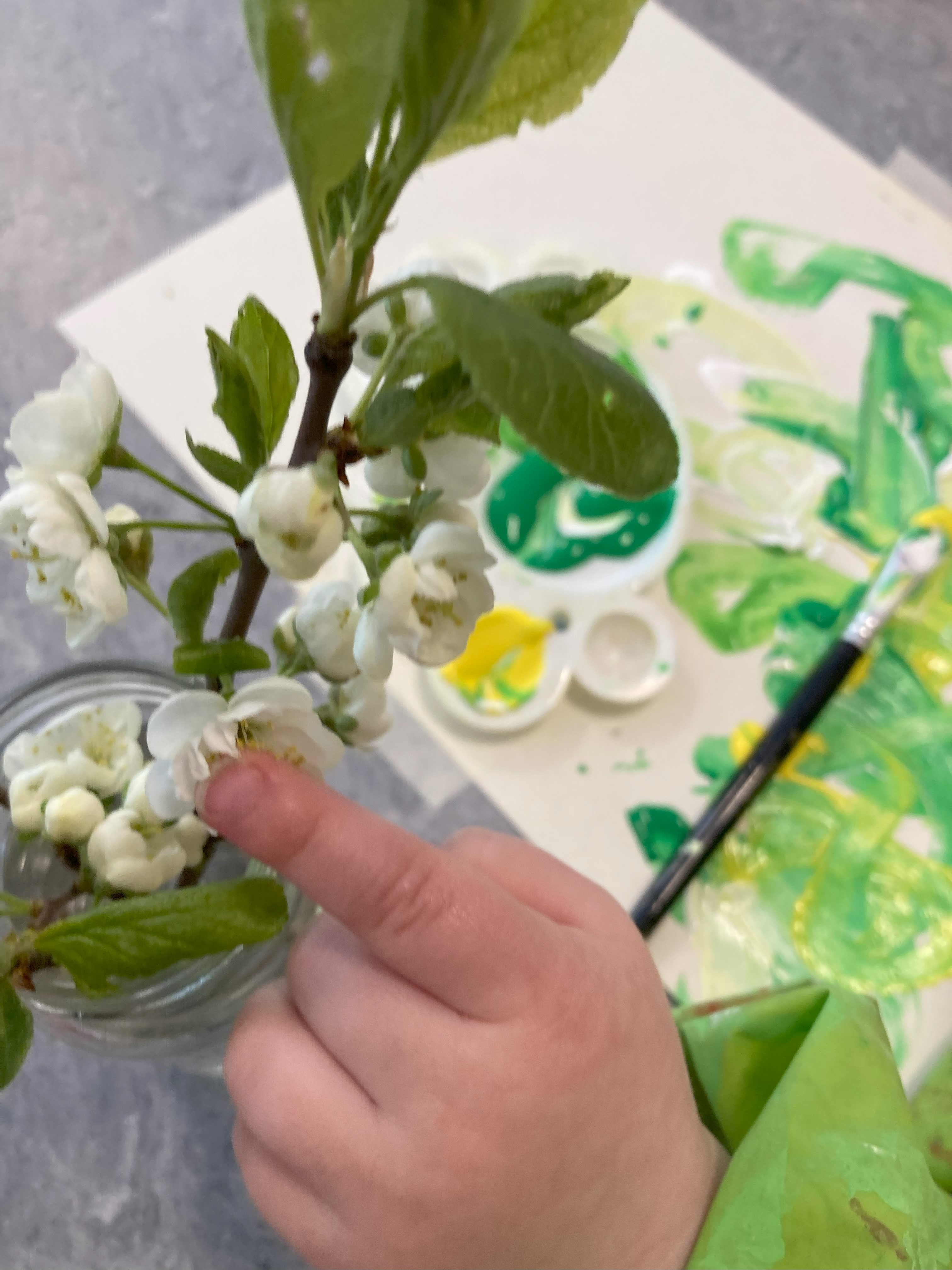 En barnhand som håller i en blomsterstängel. På bordet i bakgrunden ligger ett papper, pensel och vattenfärg. Foto. 