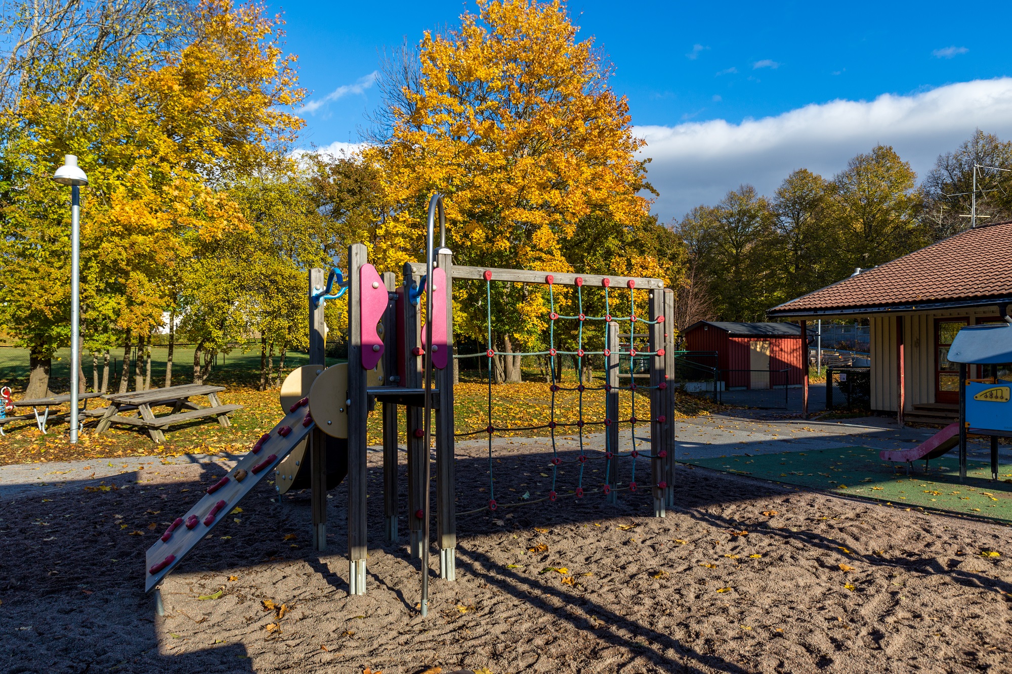 Byggnad med gård framför med fallna höstlöv, träd, sittplatser, sandlåda, klätterställning och rutschkana.