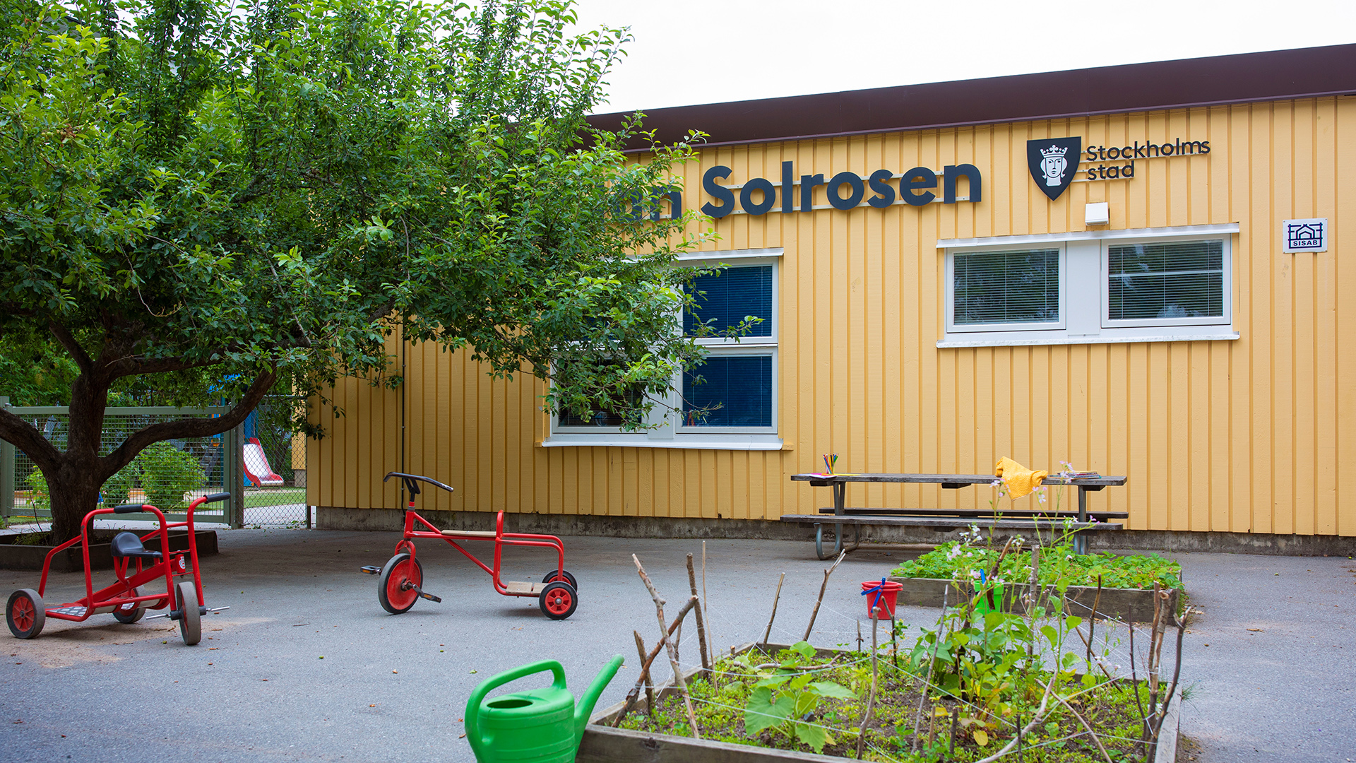 Förskolegård med träd, planteringar, gräsmatta och rutschkana. I övrigt asfalt med bord, trehjulingar och andra leksaker.