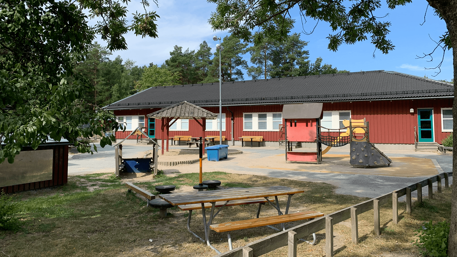 Stor gård med bänkar och lekplats. Ett enplanshus i trä i bakgrunden.