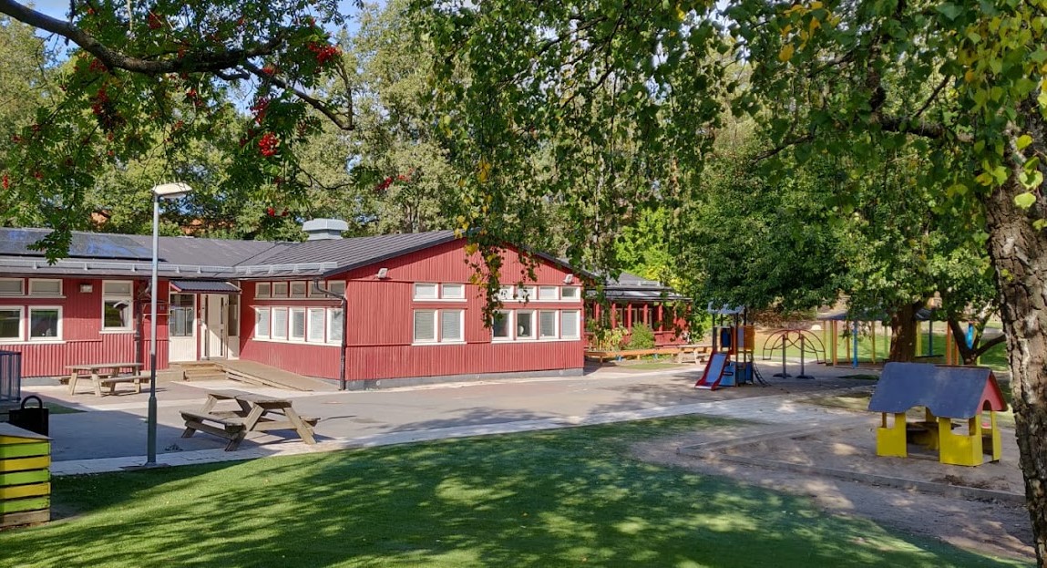 Röd låg byggnad och utemiljö med träbänkar, gult lekhus och rutschkana.