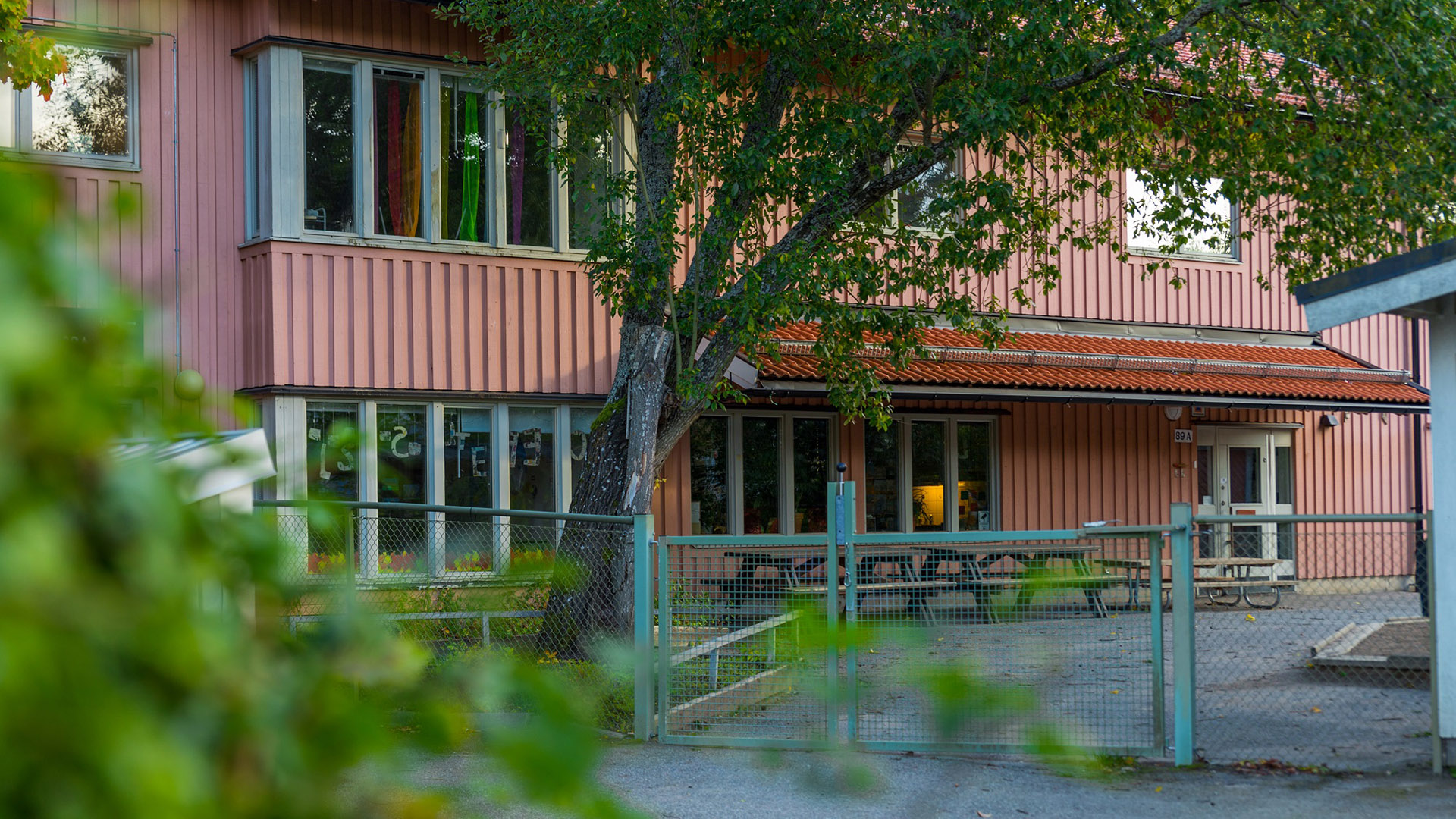 Staket och grind in till förskolebyggnad. Ett stort träd inramar entrén.