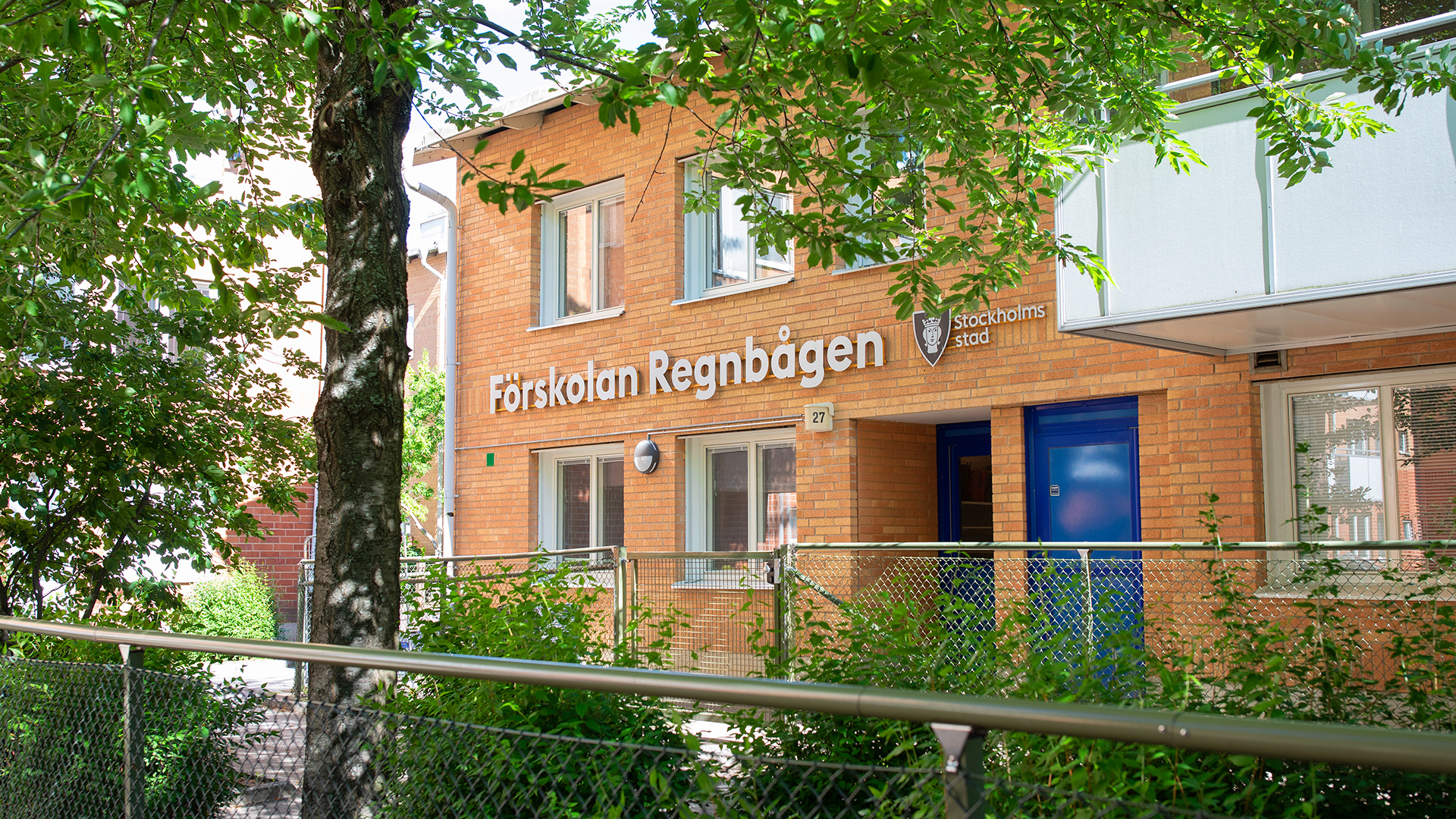 Del av förskolegård med buskar och träd. I övrigt asfalt.
