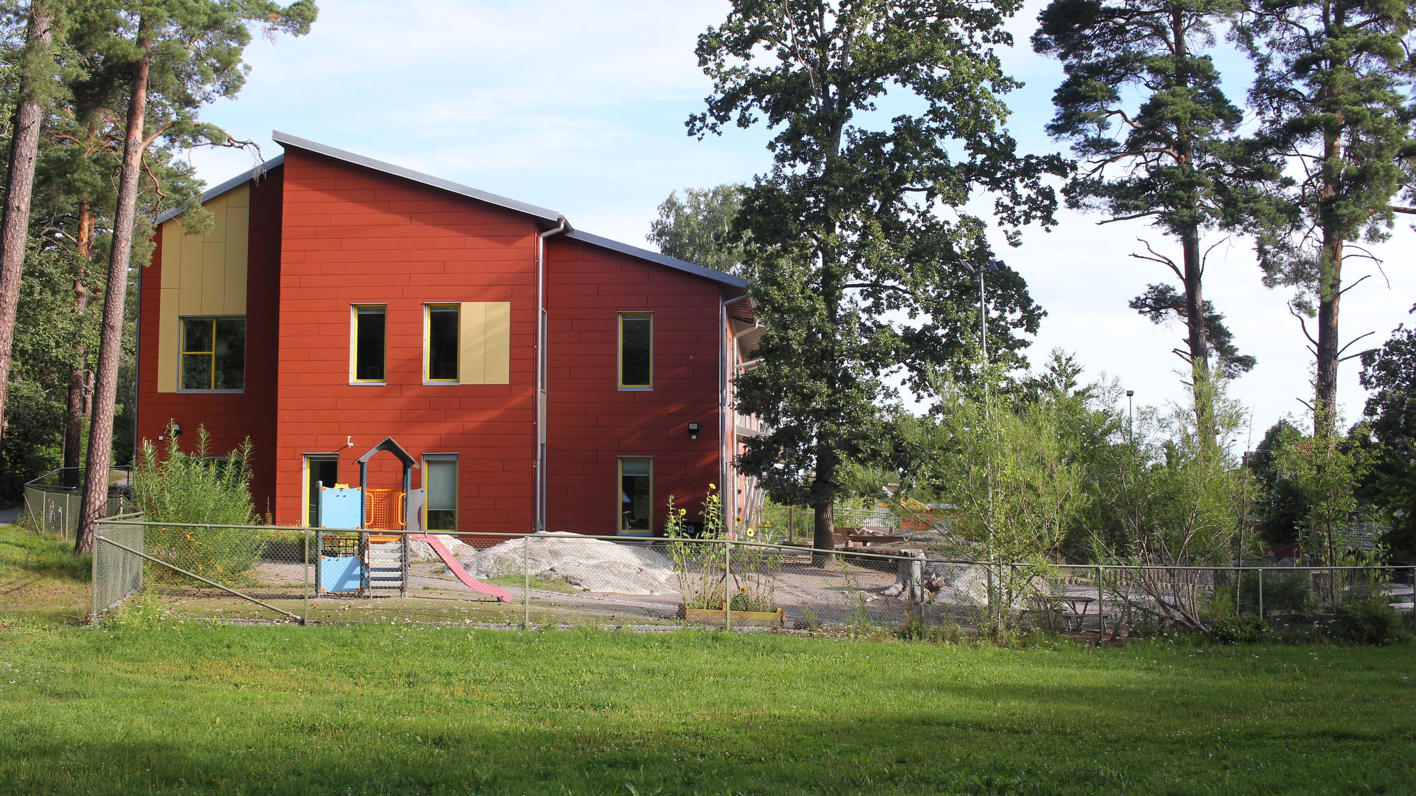Förskolan Fågelboet, ett rött tvåplanshus med gula detaljer med en stor inhägnad gård utanför. På gården syns en klätterställning och buskar och träd. Utanför är en stor gräsmatta och flera tallar.