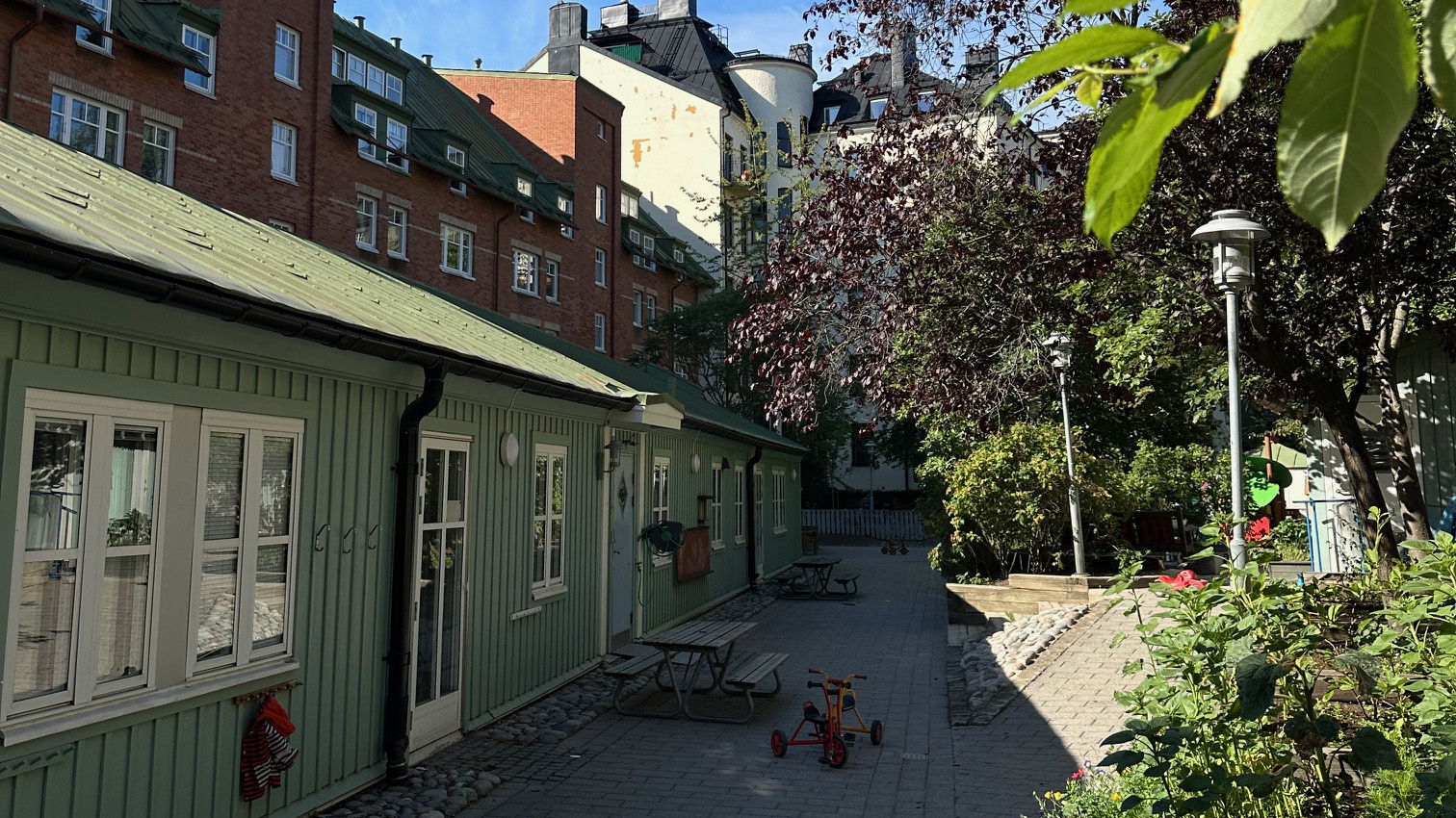 Förskola med gård framför sig med bord där man kan sitta, trehjulingar, träd och buskar.