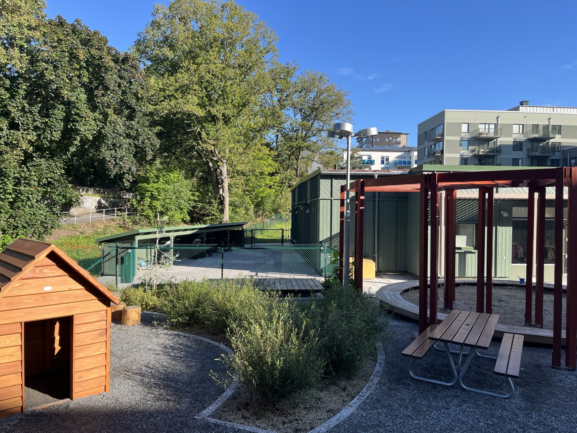 Lekyta med sittplats och minihus framför en grön förskolebyggnad. Grå flerfamiljshus syns i bakgrunden.
