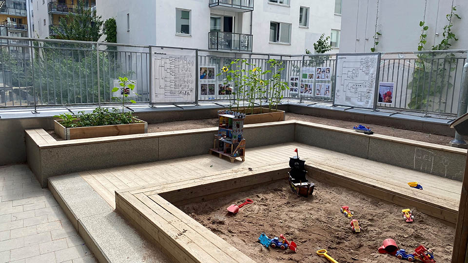 Del av gård med sandlåda, odlingslådor, och ljusa flerbostadshus i bakgrunden.