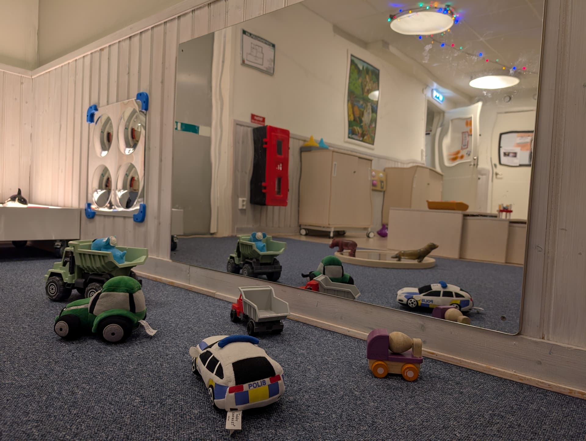 Leksaksbilar och gosedjur på golv framför spegel i lekrum, med ljusslinga i taket och möbler i bakgrunden.