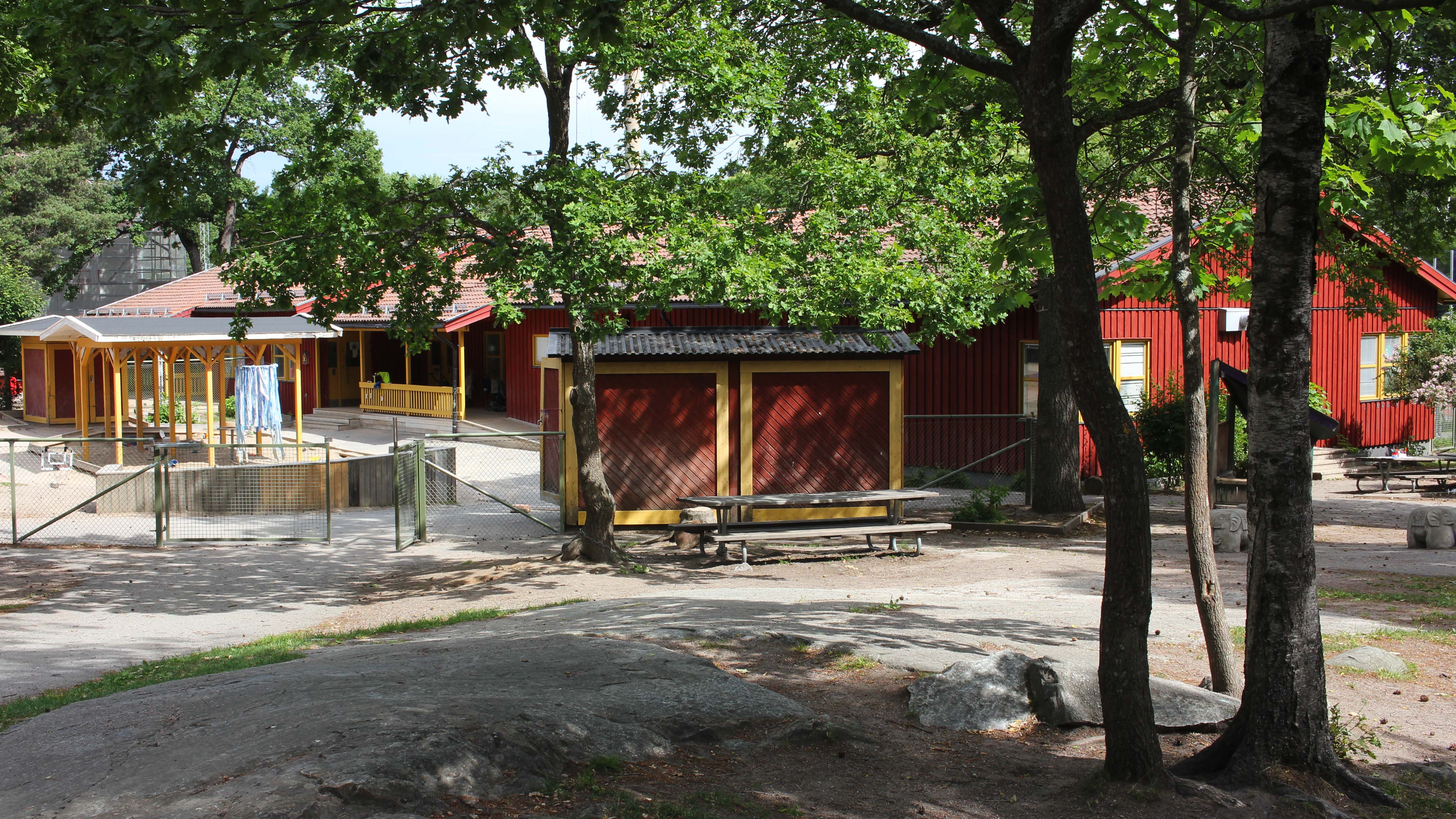 Ett rött enplanshus i trä och en inhägnad gård meden gul pergola och röda förråd intill gröna lövträd.
