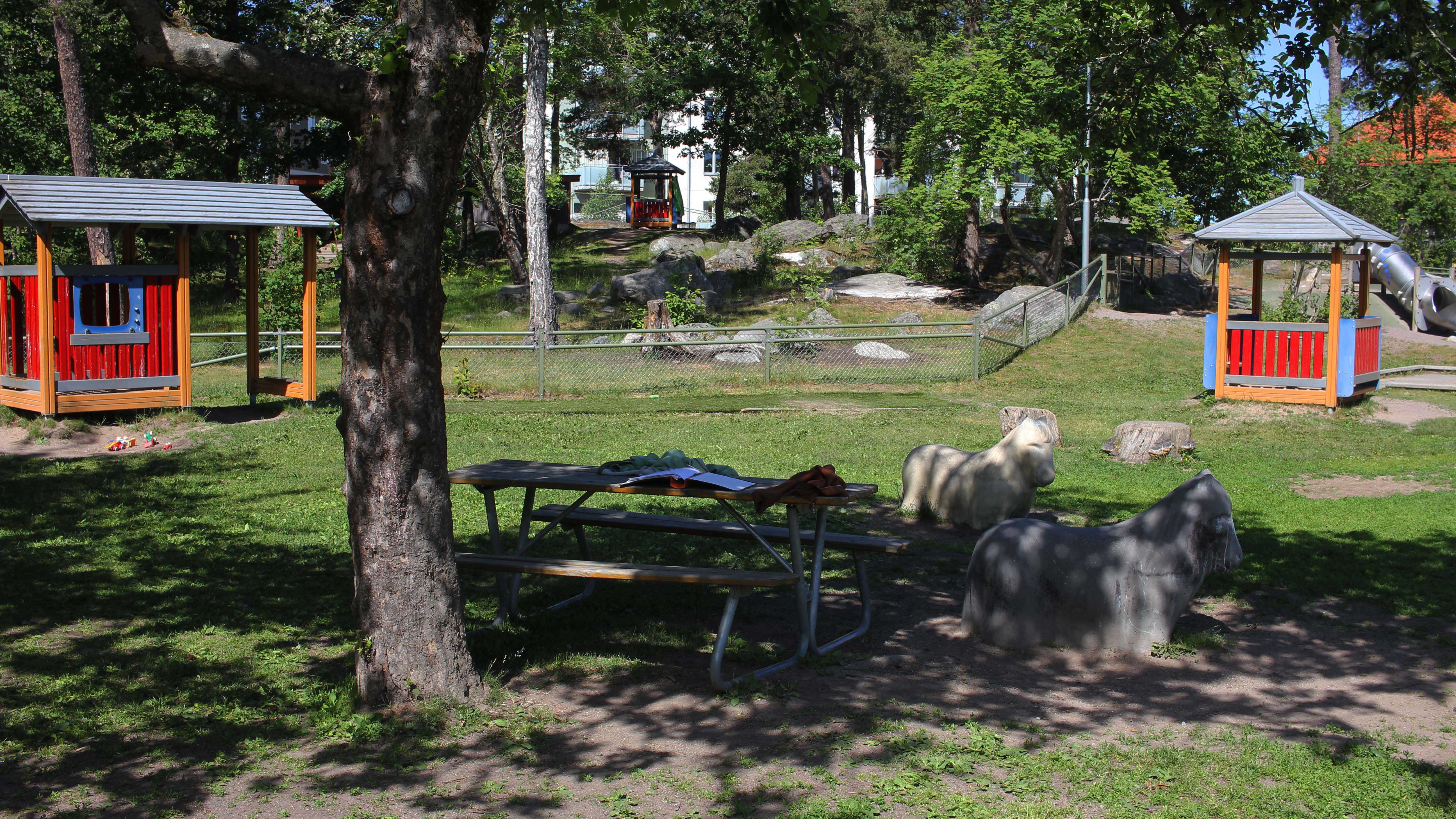 Två röda lekhus, en sittyta och konstgjorda getter på en grönskande förskolegård.
