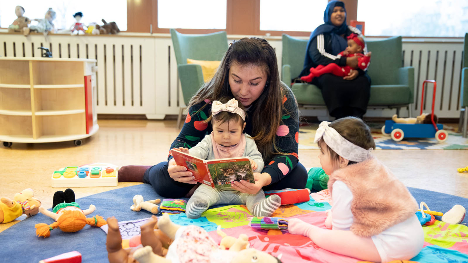 Två små barn sitter på en filt med en pedagog och tittar i en bok.