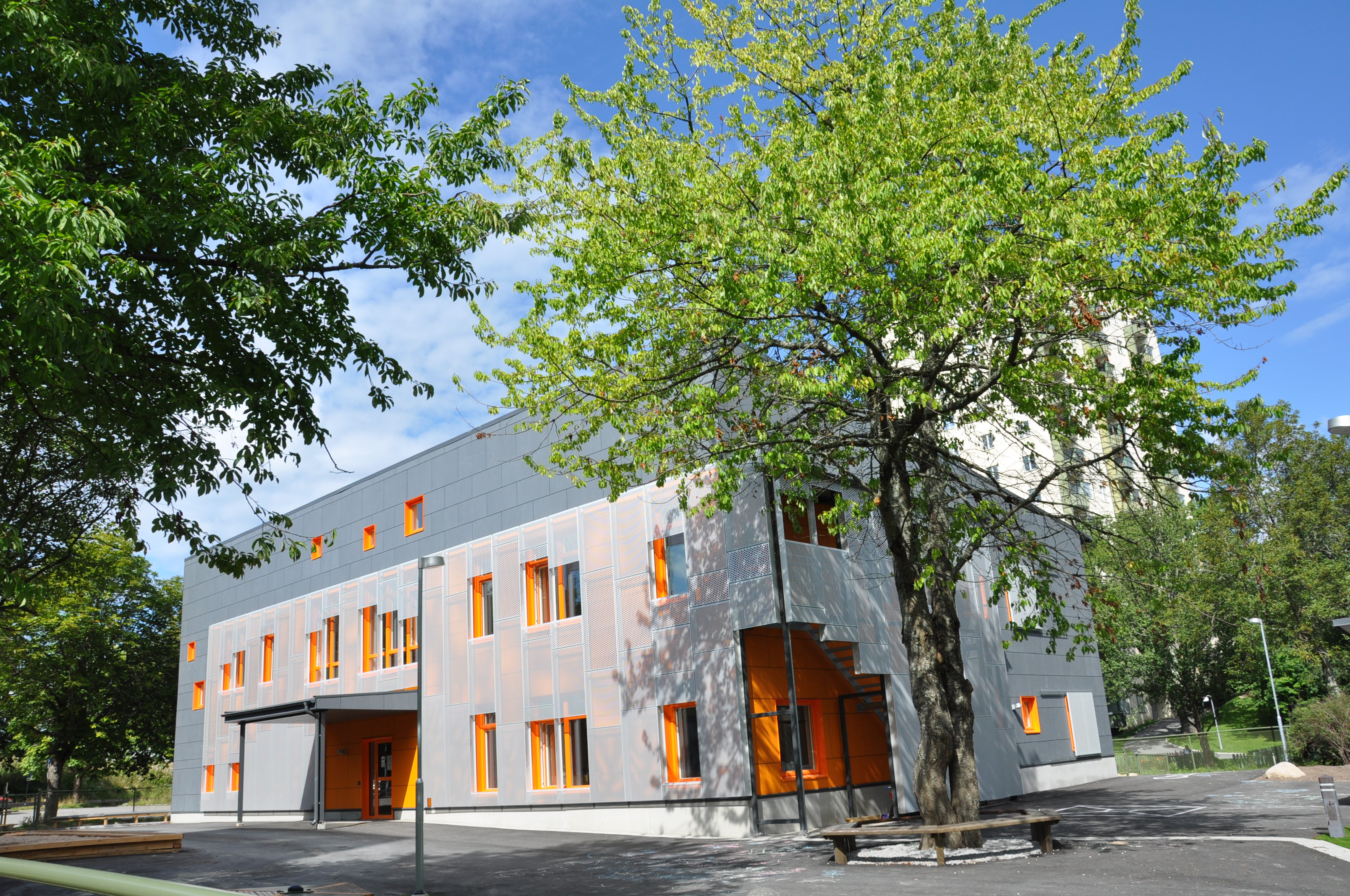 Modern tvåvåningsbyggnad, en solig gård och träd med spirande löv.