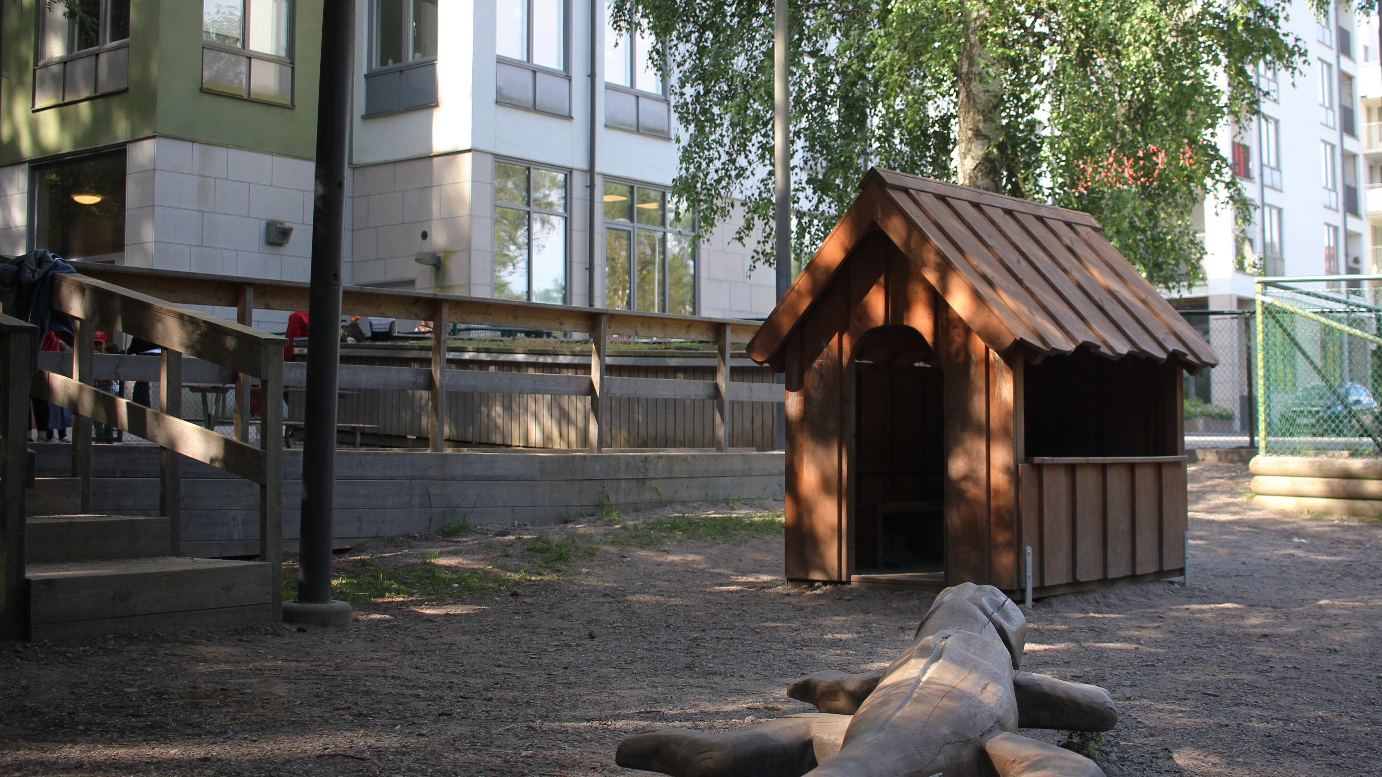 Ett lekhus i mörkt trä står på en grusad yta intill en träfigur som föreställer en krokodil. Bakom huset finns ett staket i trä och bakom staketet syns ett flerfamiljshus i vitt och grönt.
