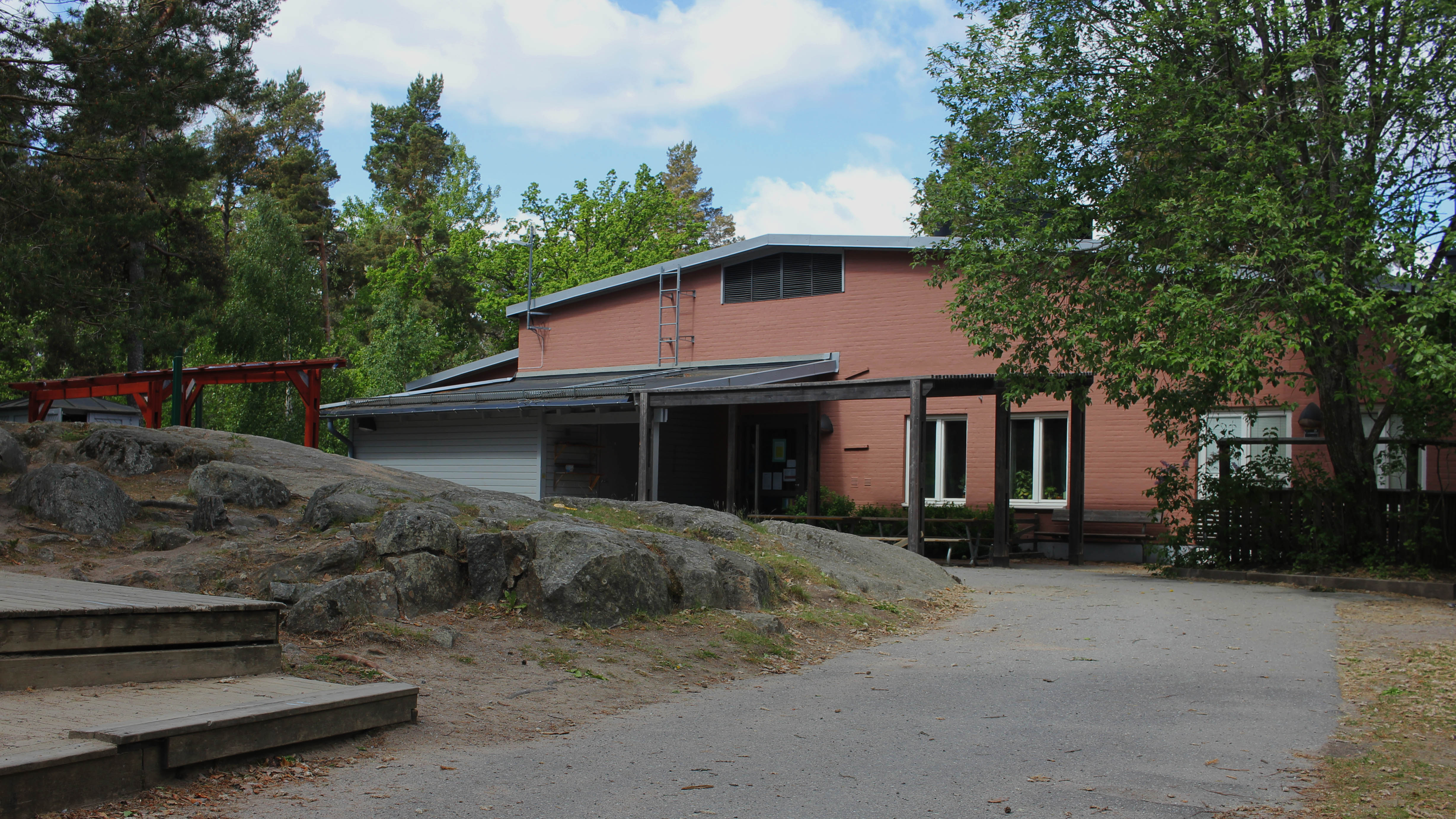 Ett rött hus i ett plan syns bland berg och träd. Intill skymtas tak till hus och en röd pergola.
