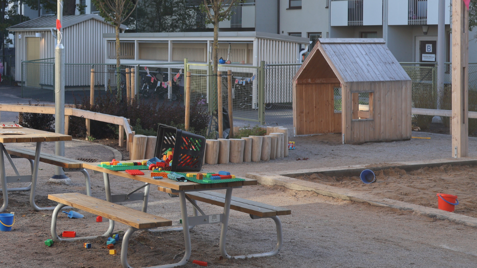 Förskolans utegård. Parkbänk med en korg med Lego på. En sandlåda. Foto. 