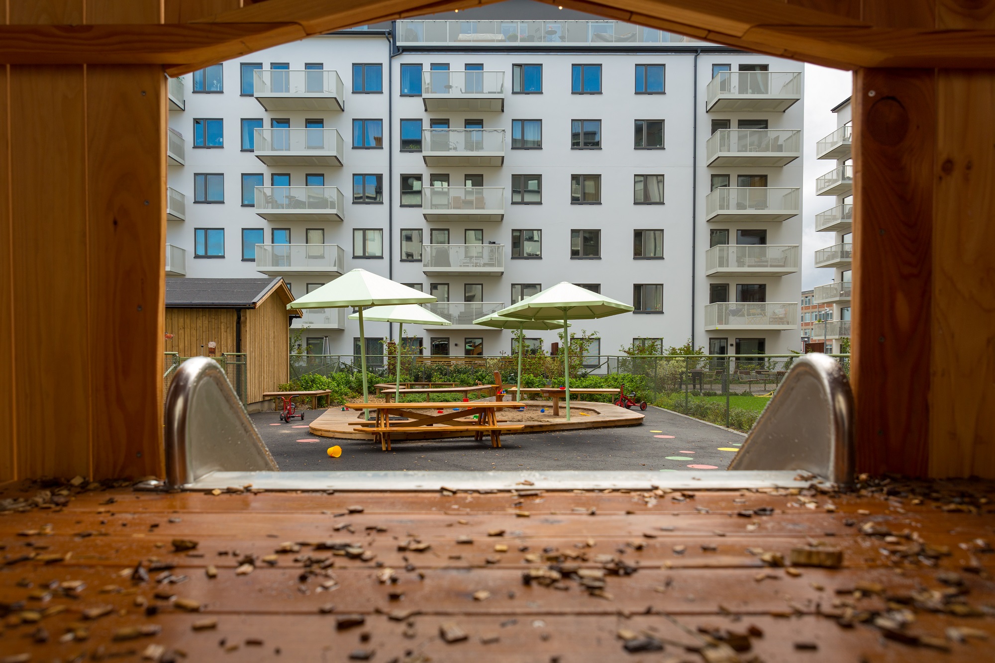 Utsikt över flerbostadshusets innergård från klätterhuset. Man ser sandlåda, lekobjekt, buskar och solskydd.