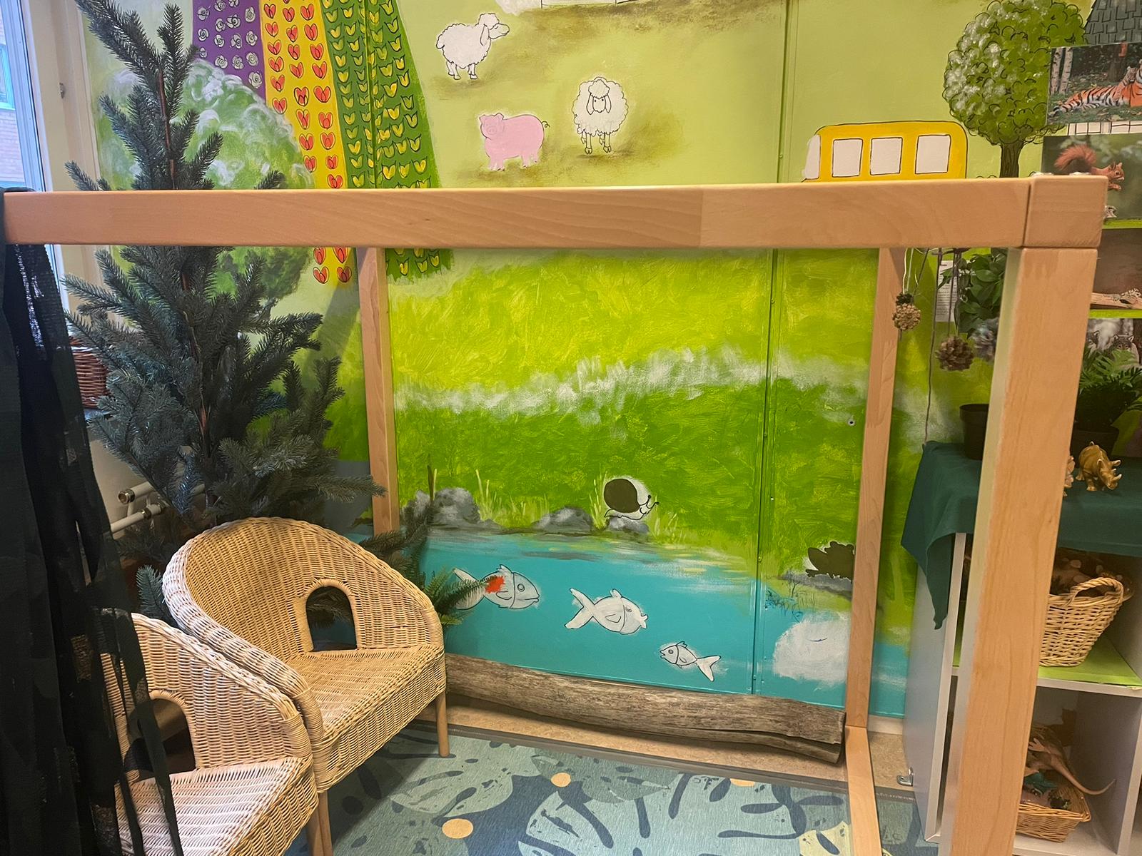 Två små stolar och hyllor med plastdjur som står framför en väggmålning med natur, djur och fiskar i en fiskdamm.