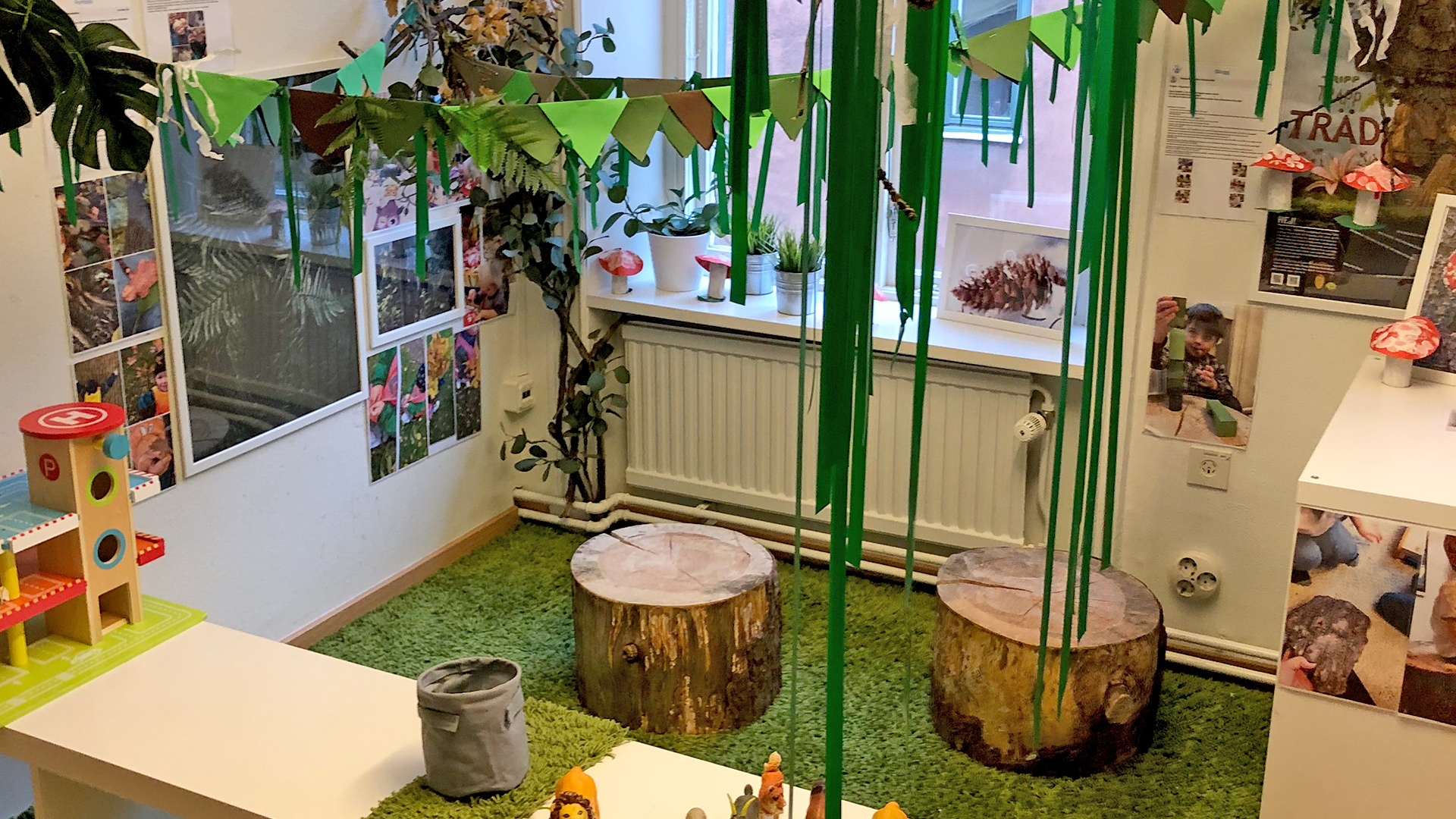 Lekrum på förskola med en stor matta för lek och hyllor för leksaker. Bilbana står på ett skrivbord och två stubbar står på en grön fransig matta.