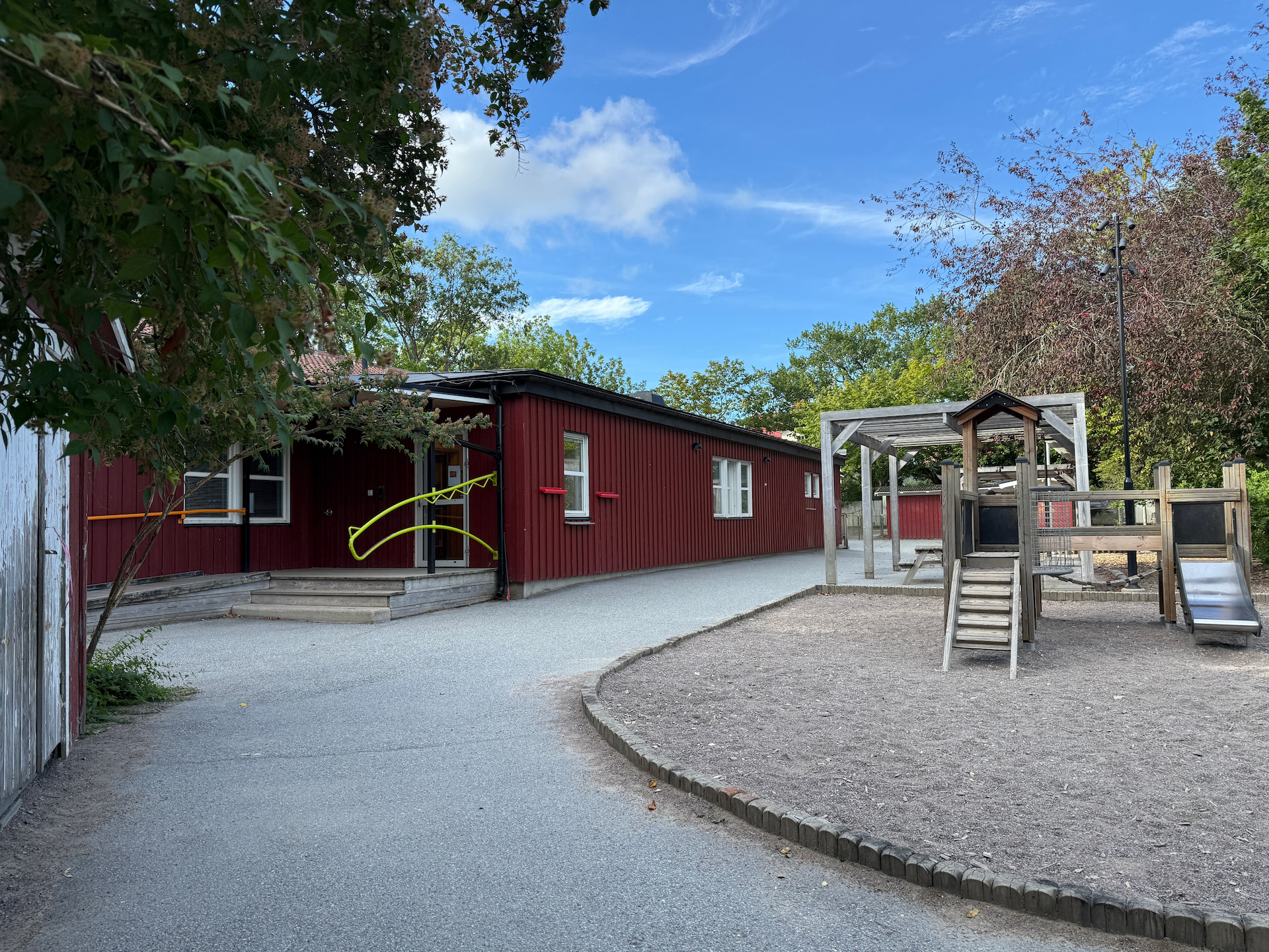 Röd förskolebyggnad med en asfalterad gång och en lekplats med klätterställning och rutschkana.