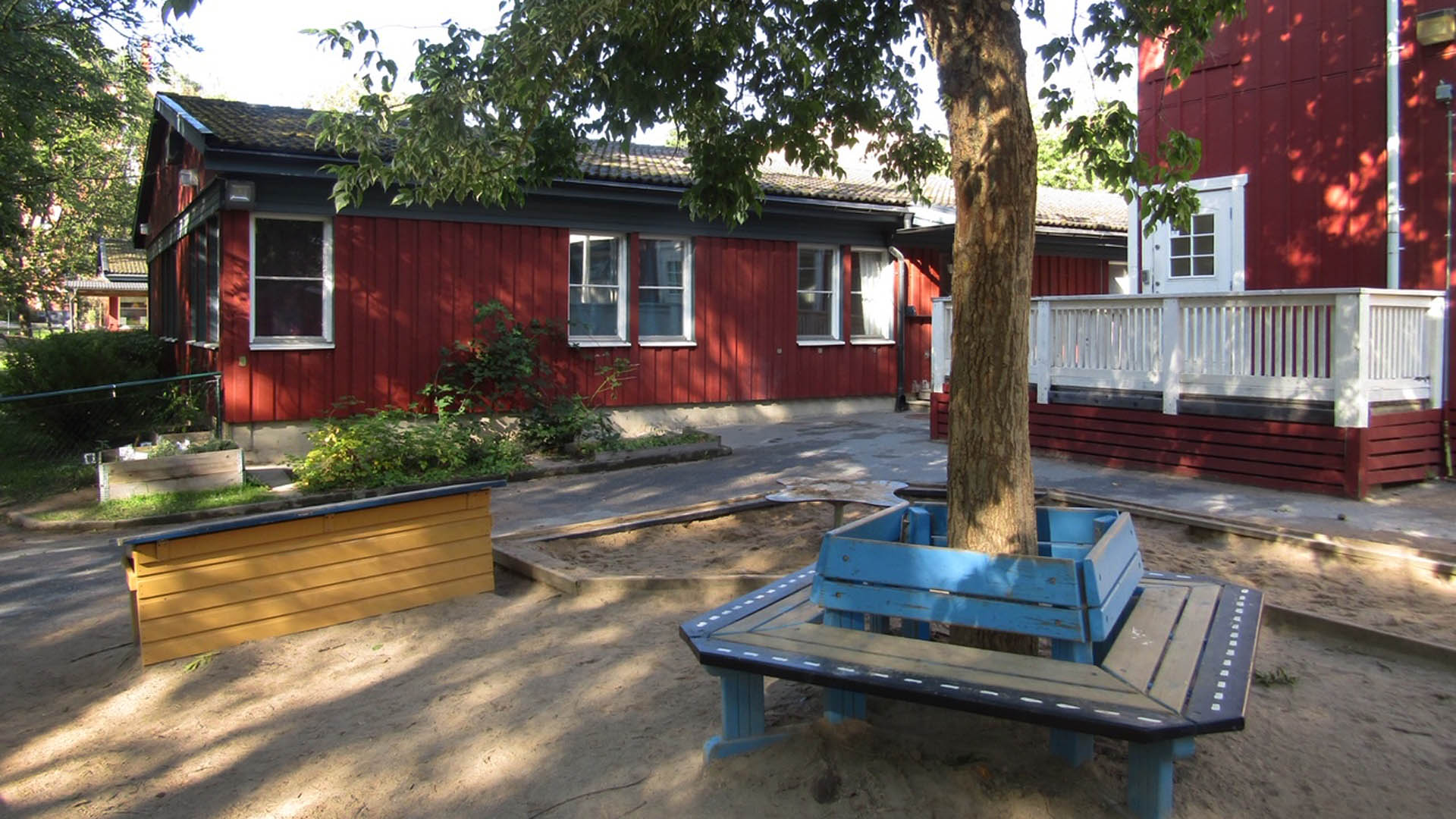 Röda träfasader och lekmiljö med sandlåda och träd i förgrunden.