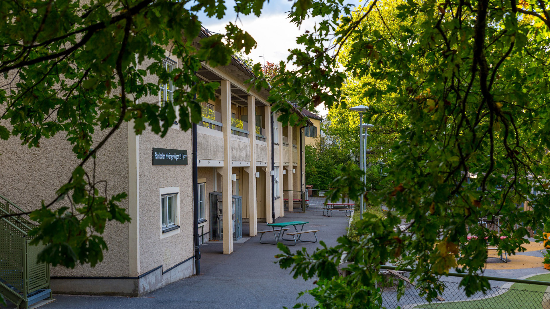 Förskolebyggnad med asfalterad gårdsplan skymtar mellan trädgrenar. Sittplatser vid entrén.