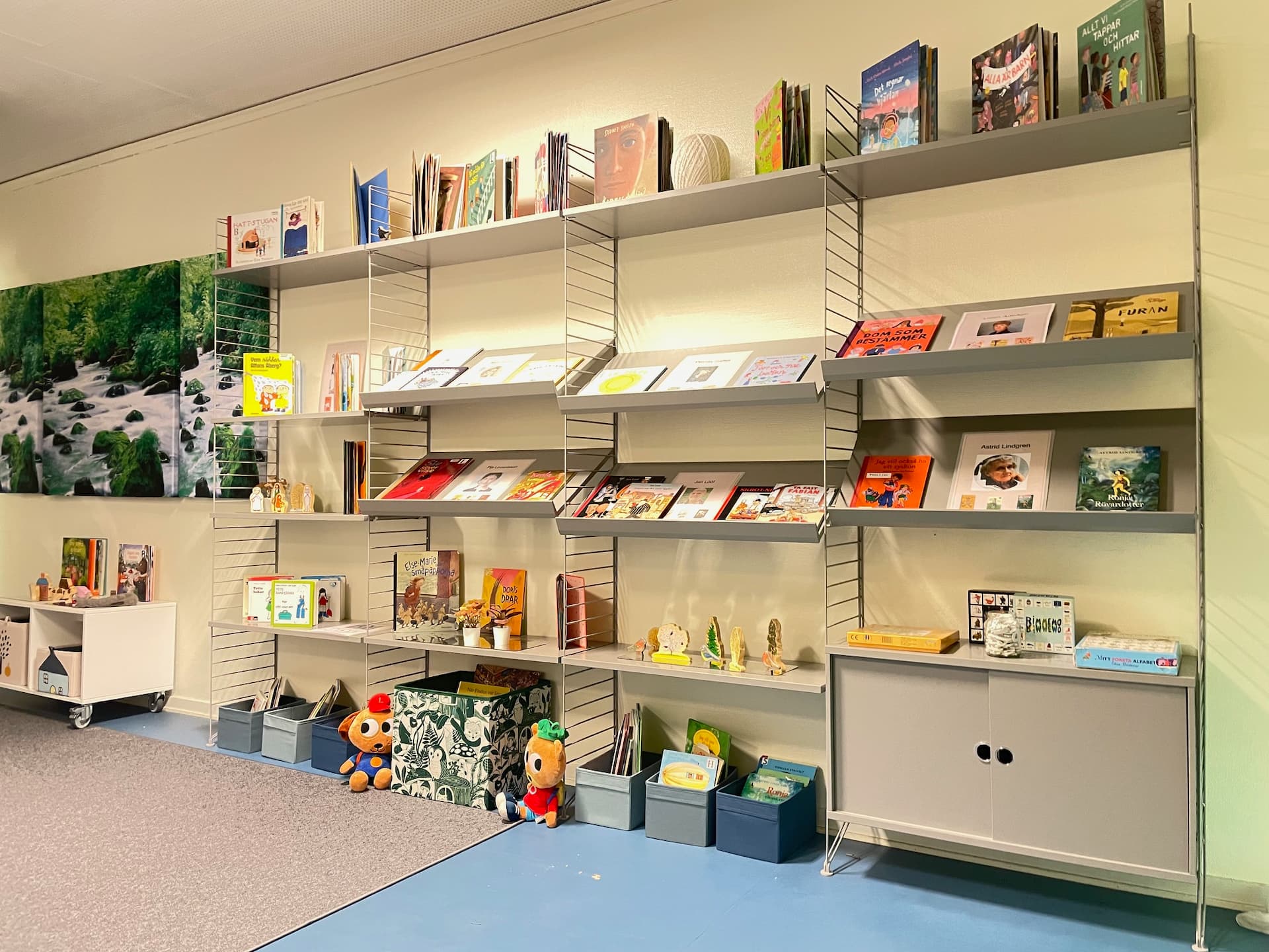 Barnböcker visas på grå vägghyllor i ett bibliotek med lådor, spel och mjukisdjur på golvet.