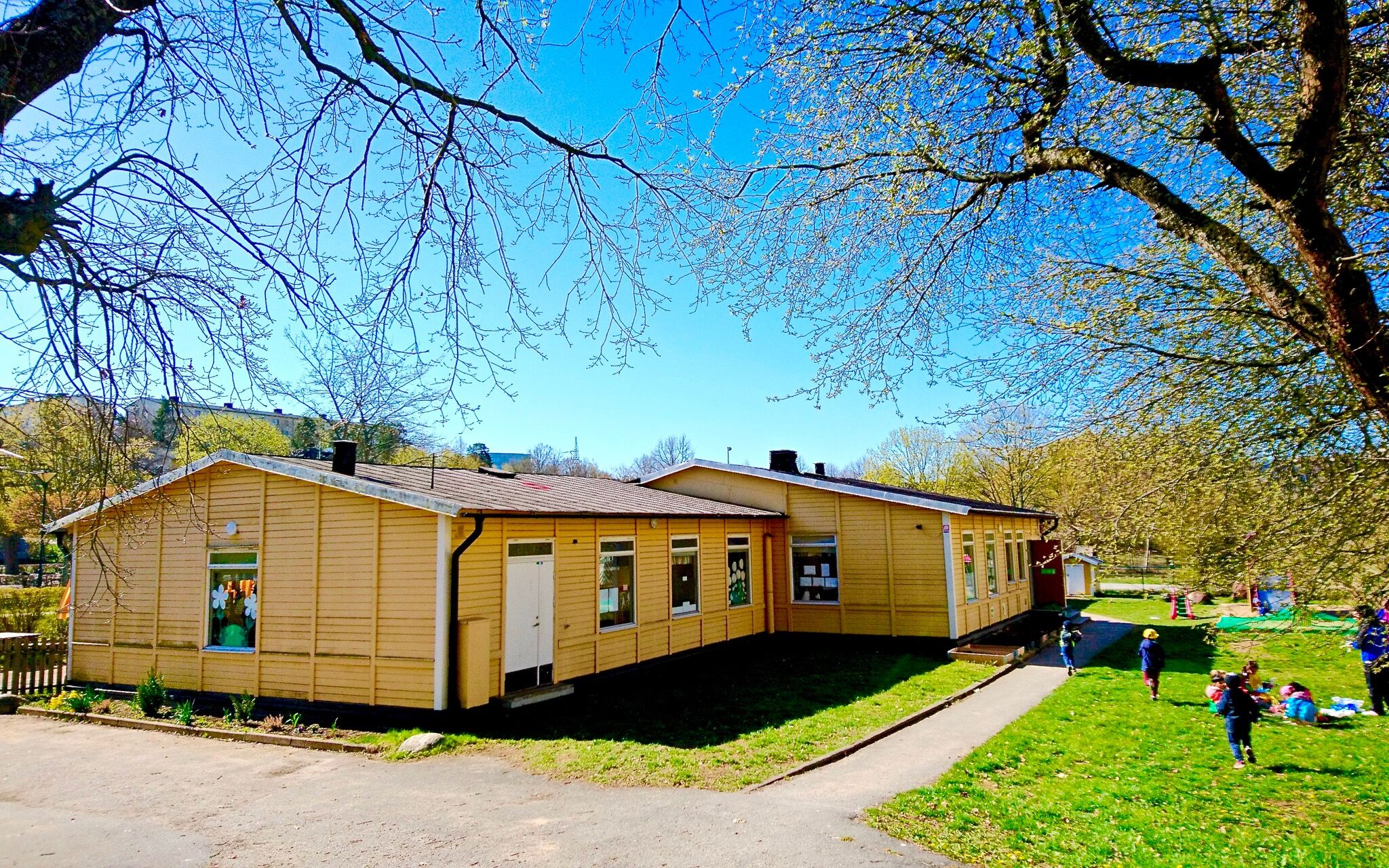 Förskola, gult hus, blå himmel, barn som leker på gräset.